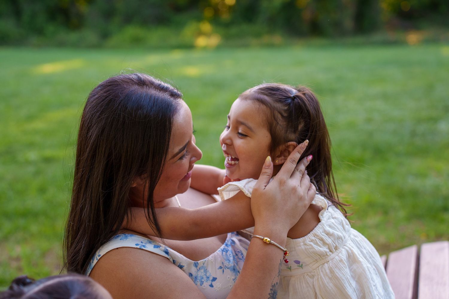 A mother and young daughter share a joyful moment outdoors, smiling at each other in a lush green backyard.