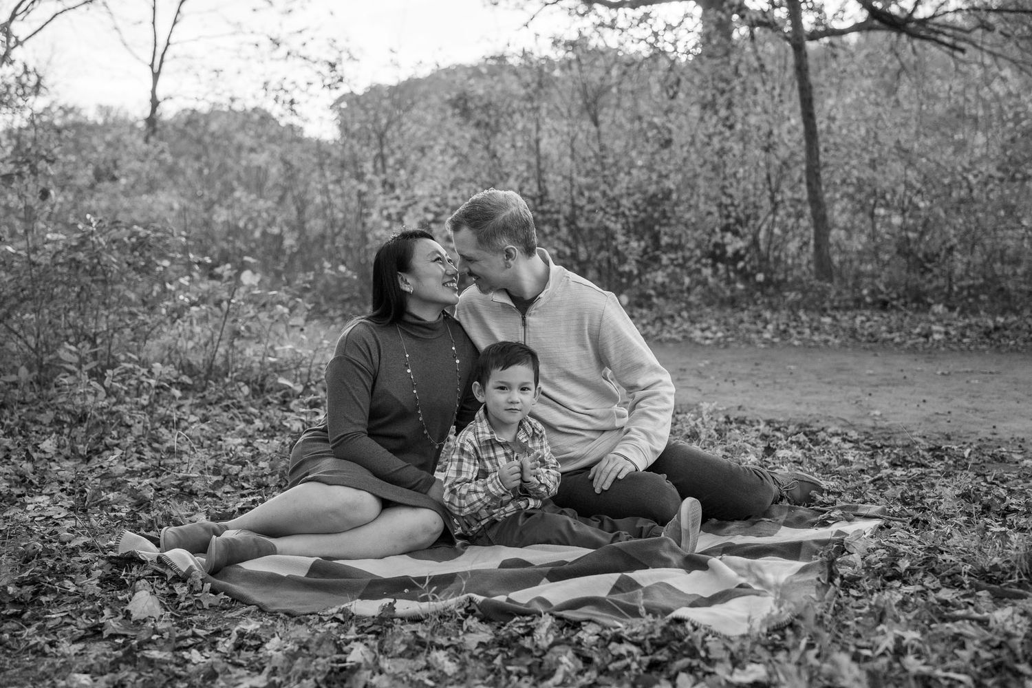Family relaxing on blanket among fallen leaves during black and white outdoor photo session.