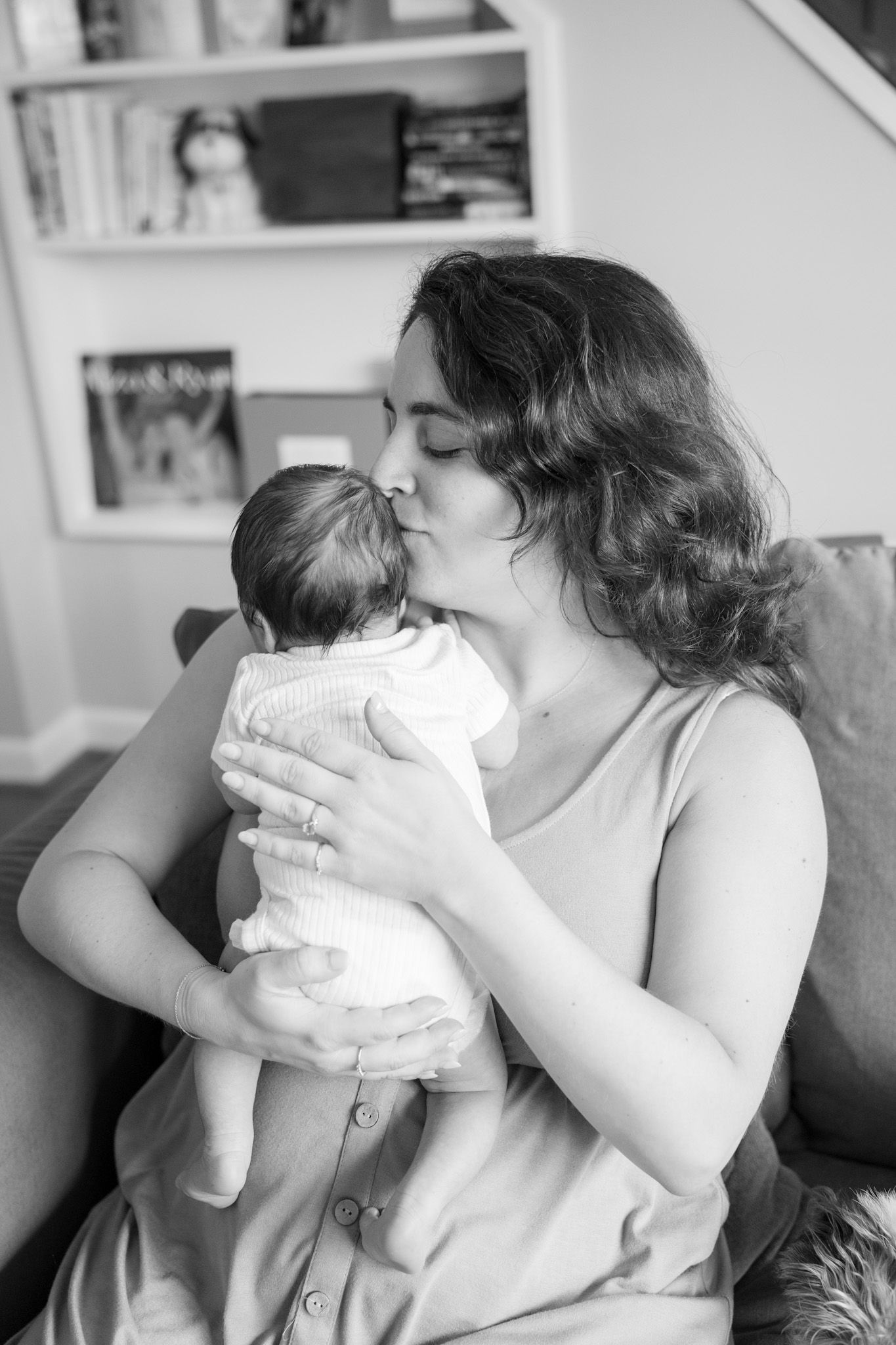 Black and white photo of a mother kissing her newborn baby while sitting on a couch at home.