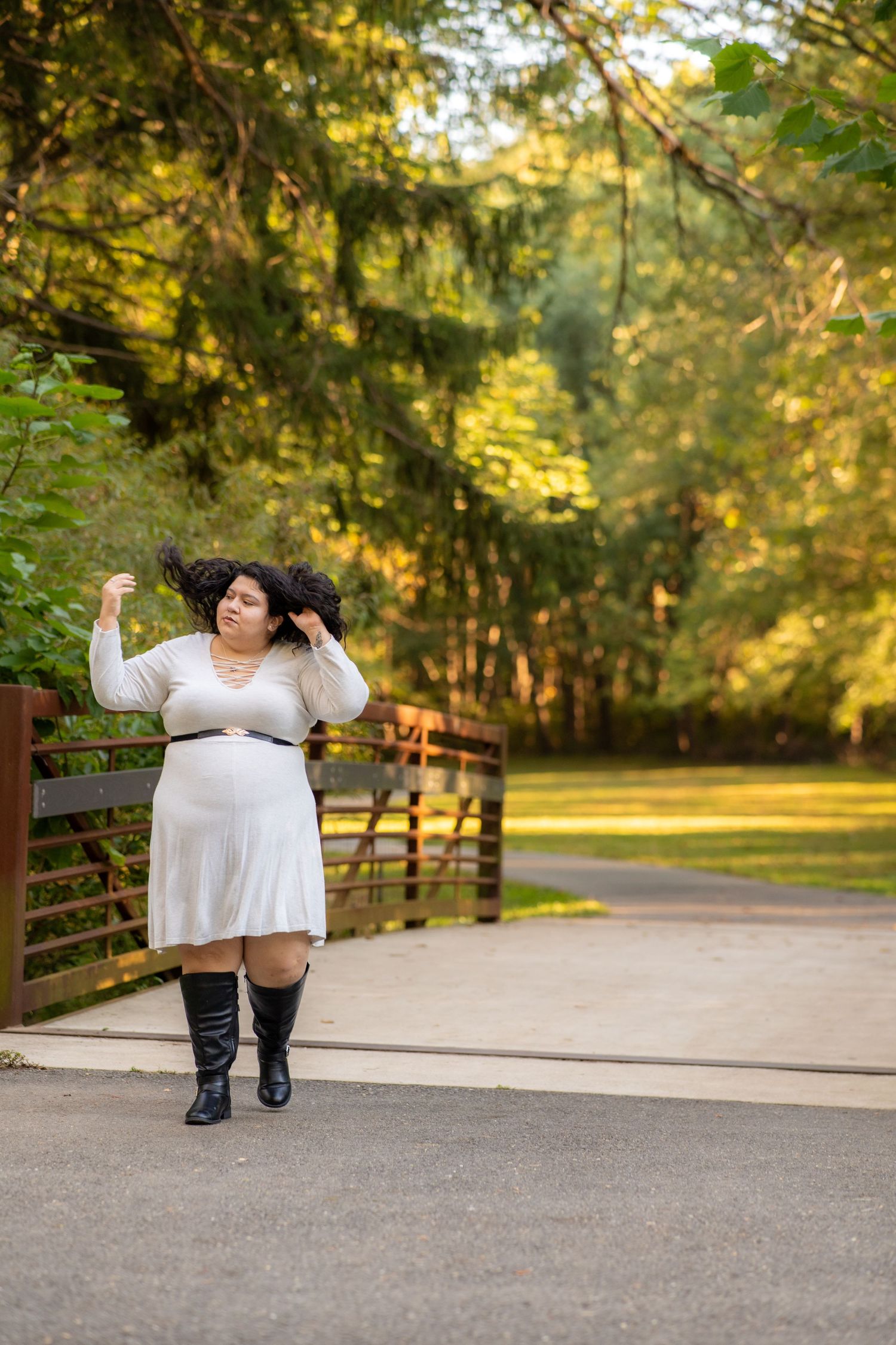A figure in a white flowing dress and boots poses playfully on a paved path surrounded by autumn trees and fencing.