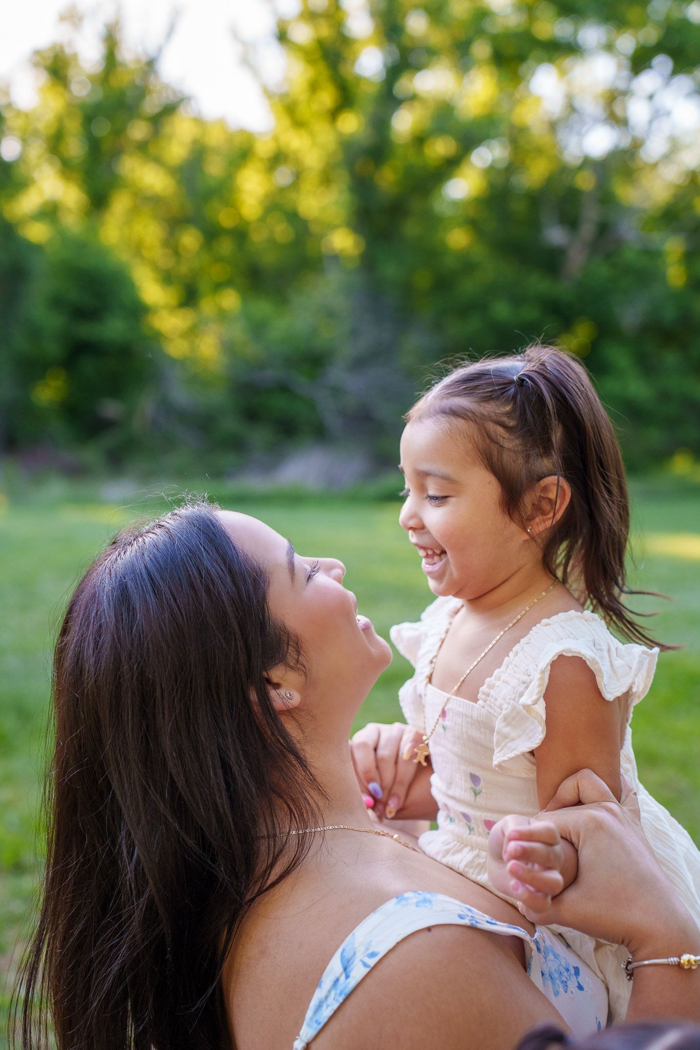 A joyful mother lifts her laughing toddler daughter outdoors in a sunny green park setting.