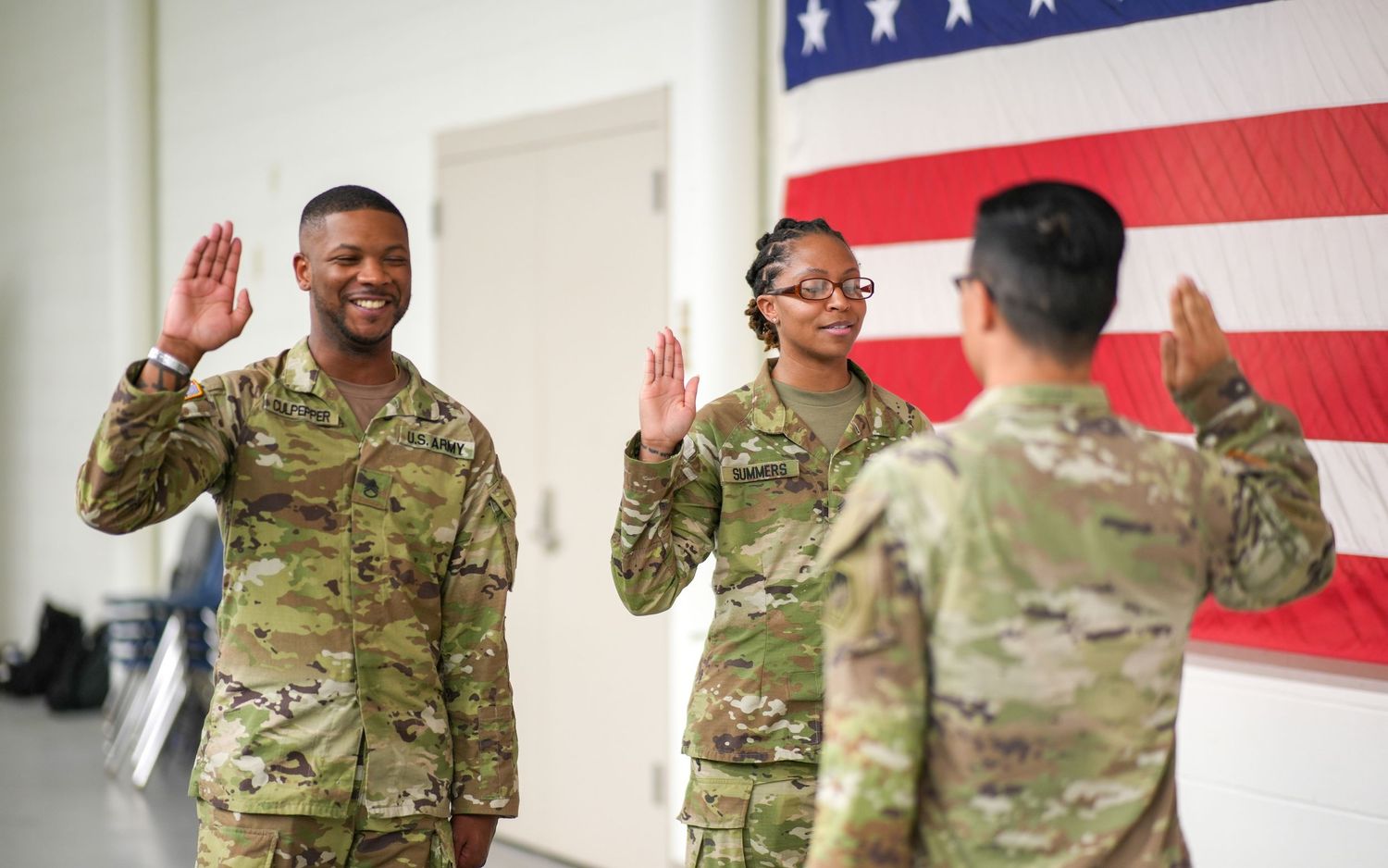 Military personnel raise their hands during an oath ceremony against American flag background.