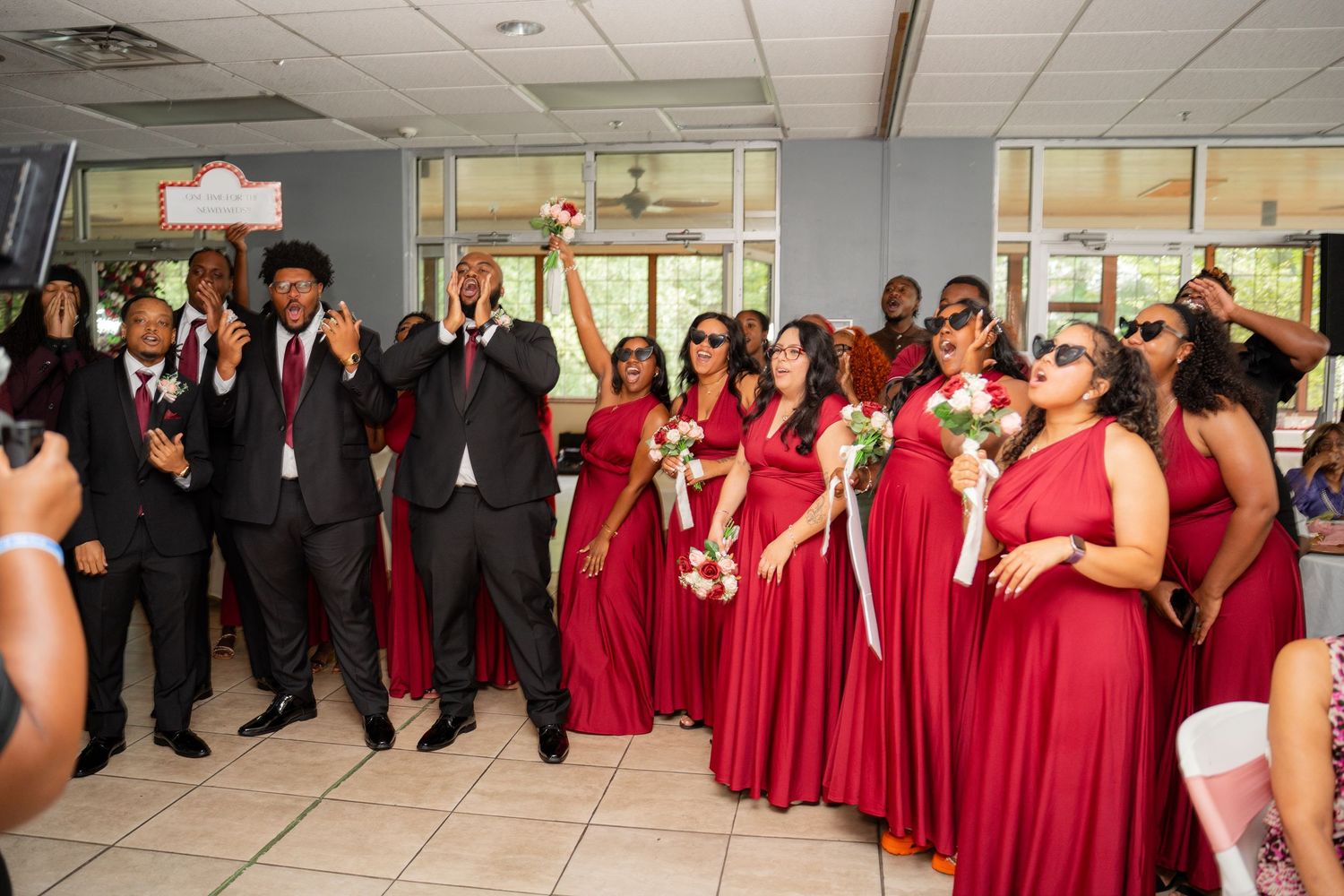 Wedding party in red formal dresses and black suits posing in reception hall.