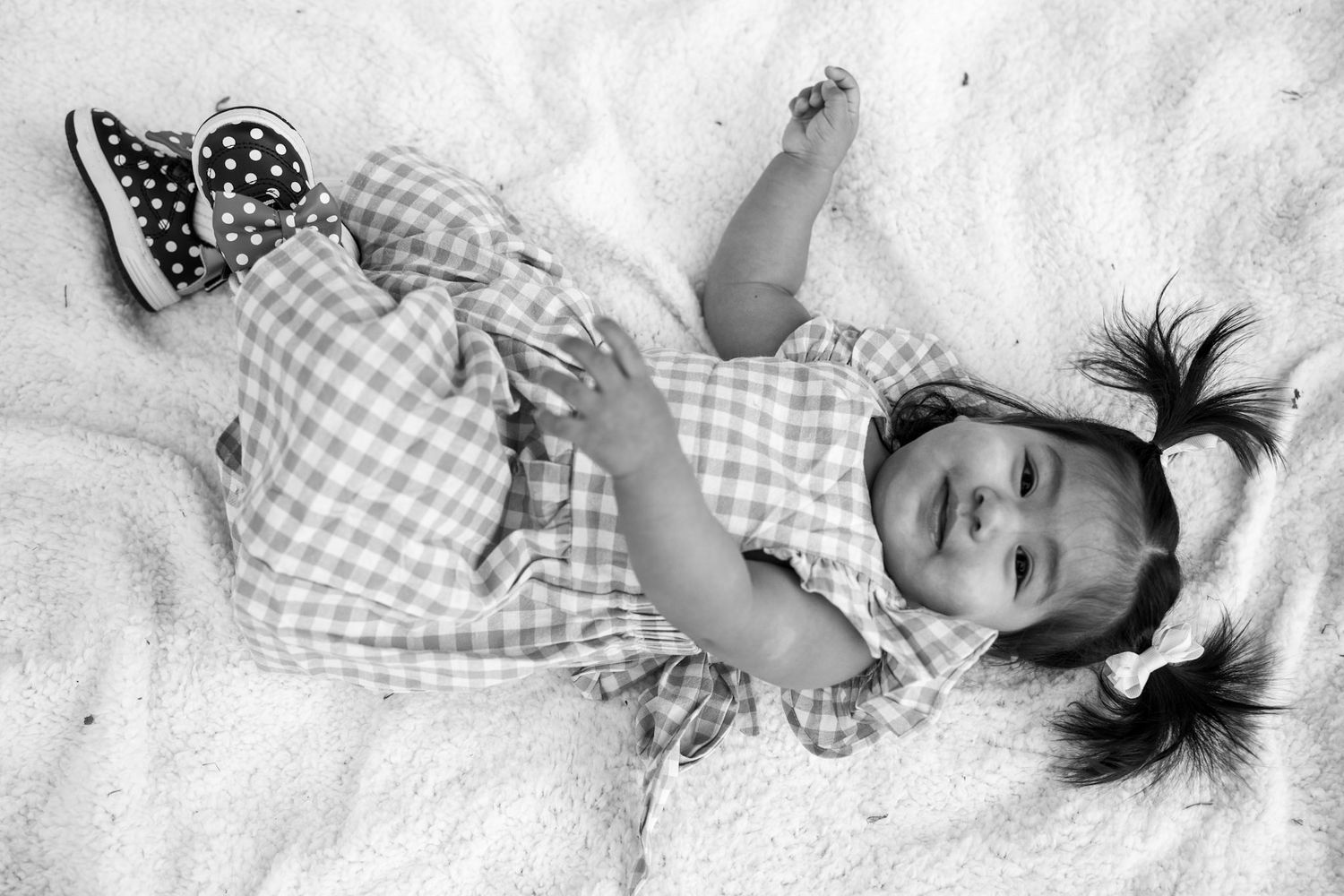 Black and white photo of a cute baby girl in gingham dress with pigtails, lying on a fluffy white blanket.