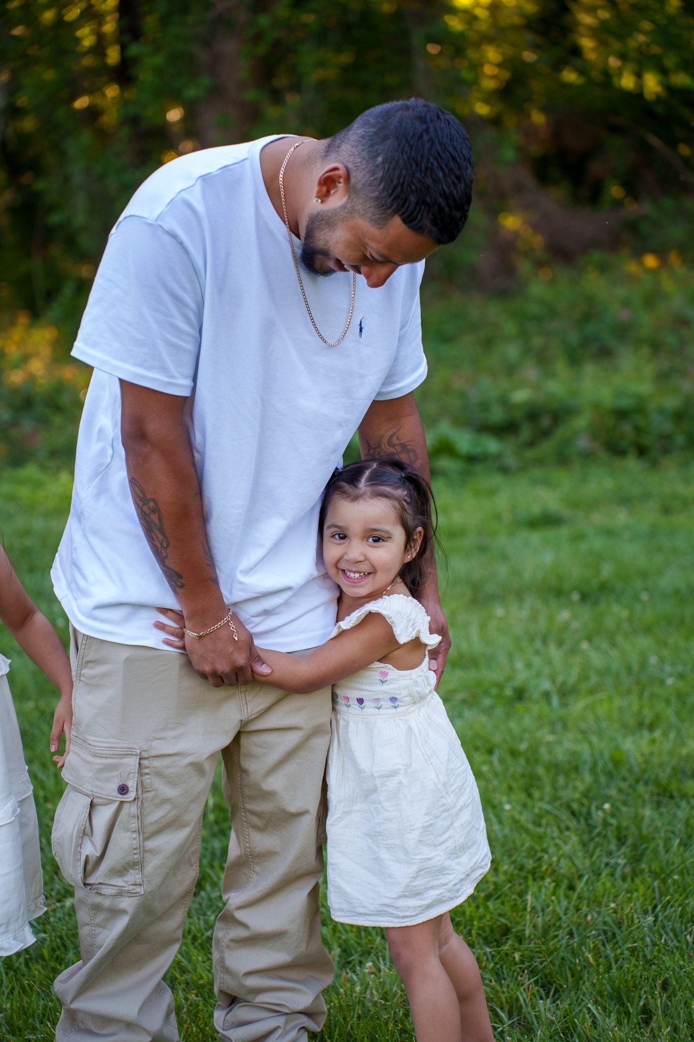 A father in a white shirt and khaki pants shares a warm hug with his smiling young daughter in a white dress outdoors.