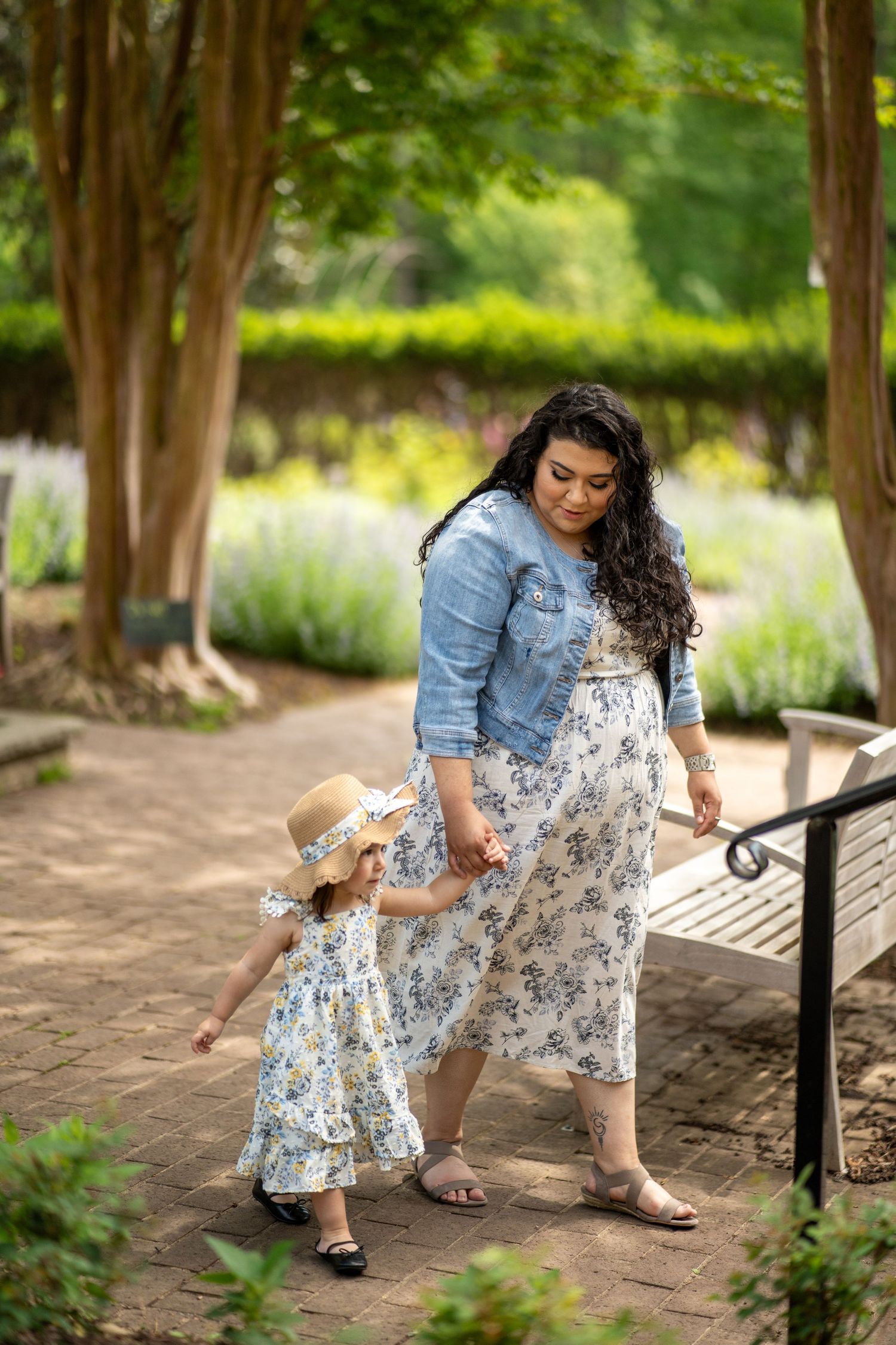 A parent and child in matching floral dresses and denim jackets walk together on a garden path surrounded by greenery.