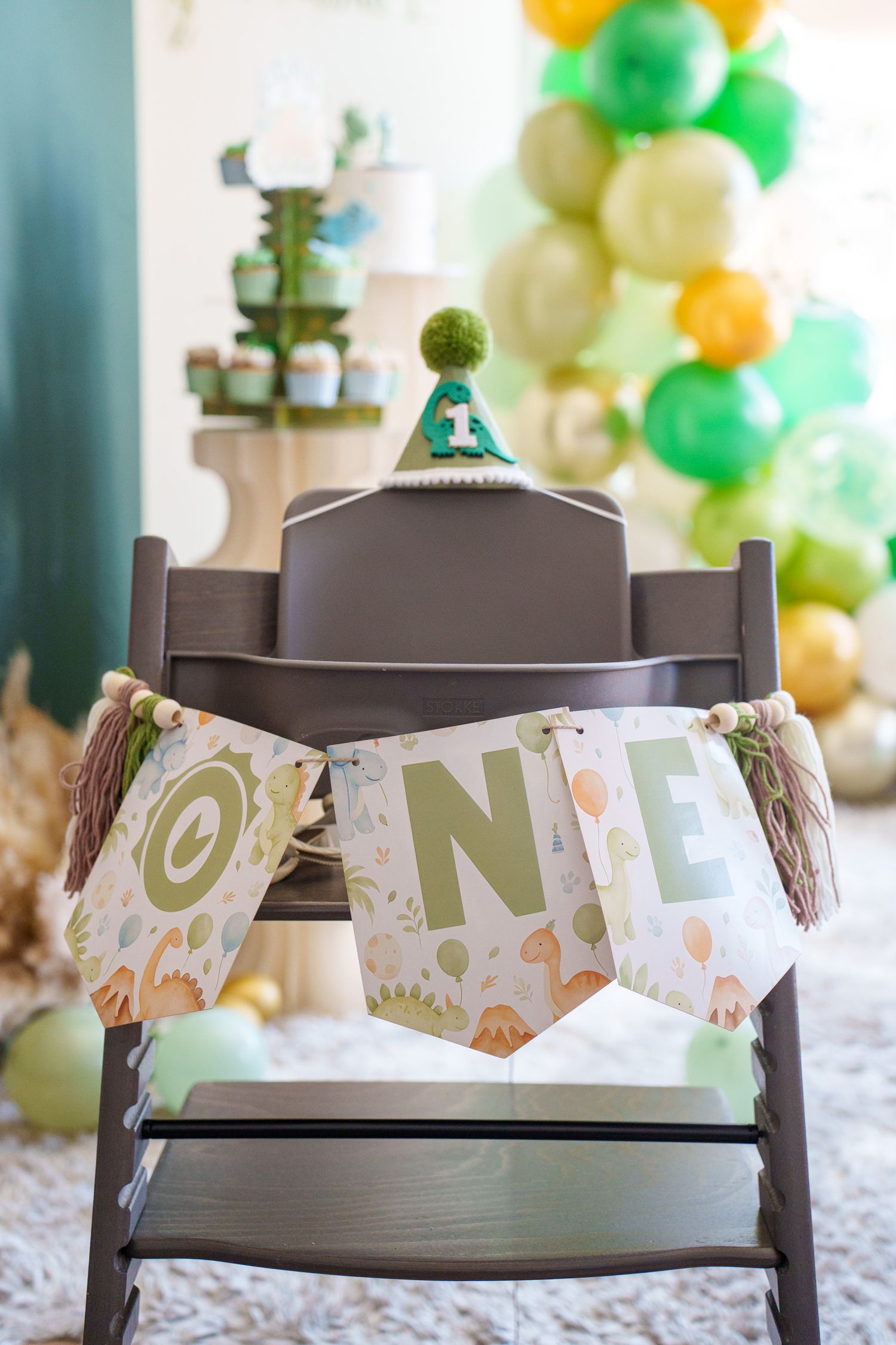A festive high chair decorated with a banner and green and gold balloons for a first birthday celebration.