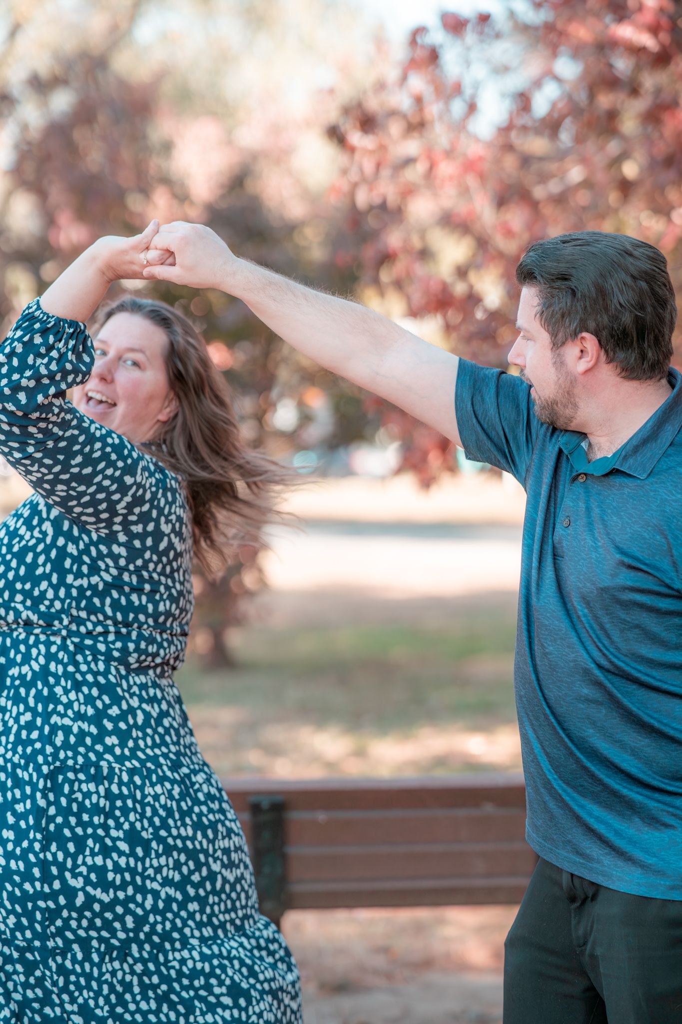 A playful couple dancing and twirling together outdoors against a backdrop of autumn trees.