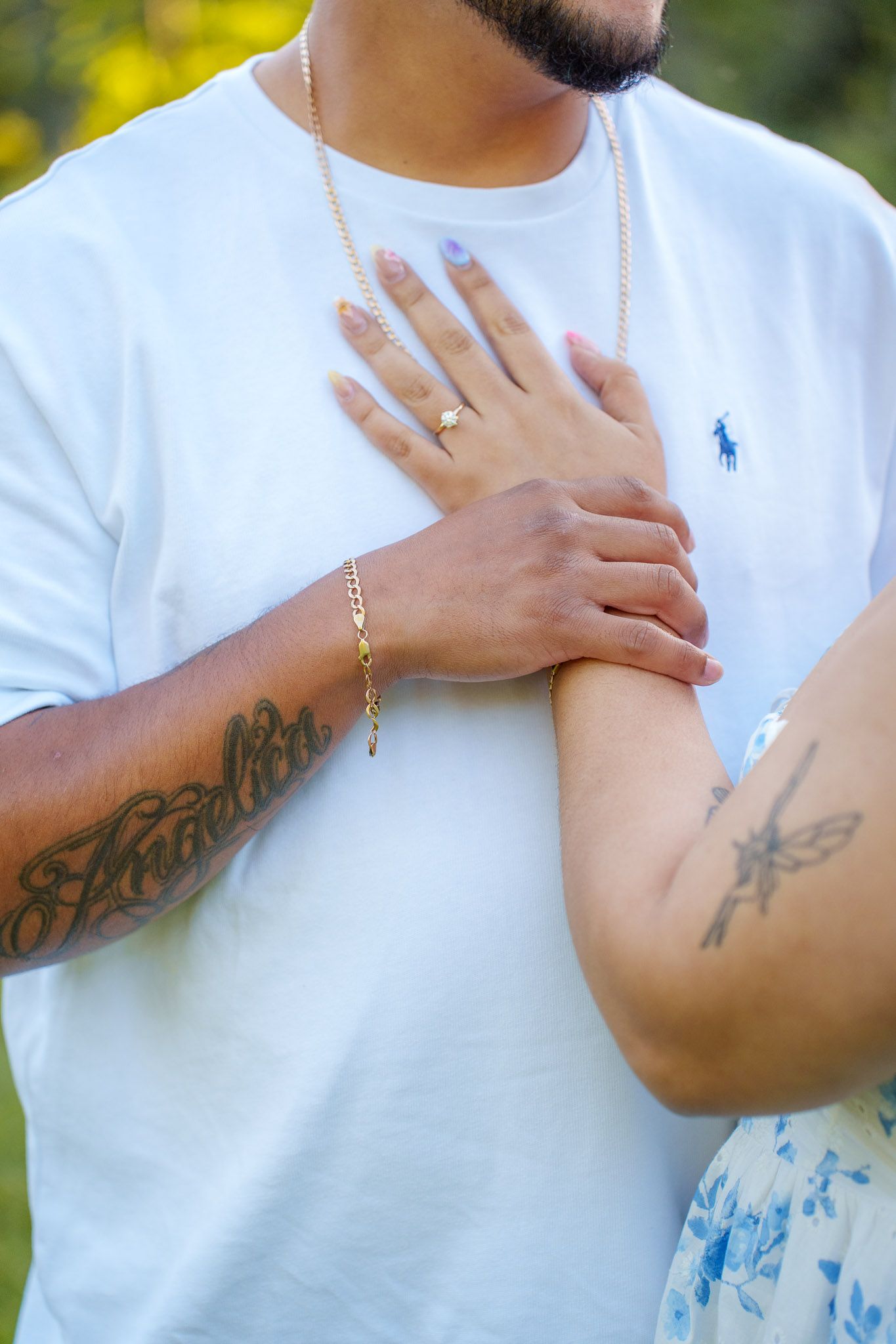 Couple holding hands showing gold bracelet, ring, blue nails, and tattoos, man wearing white Polo Ralph Lauren tee and chain.