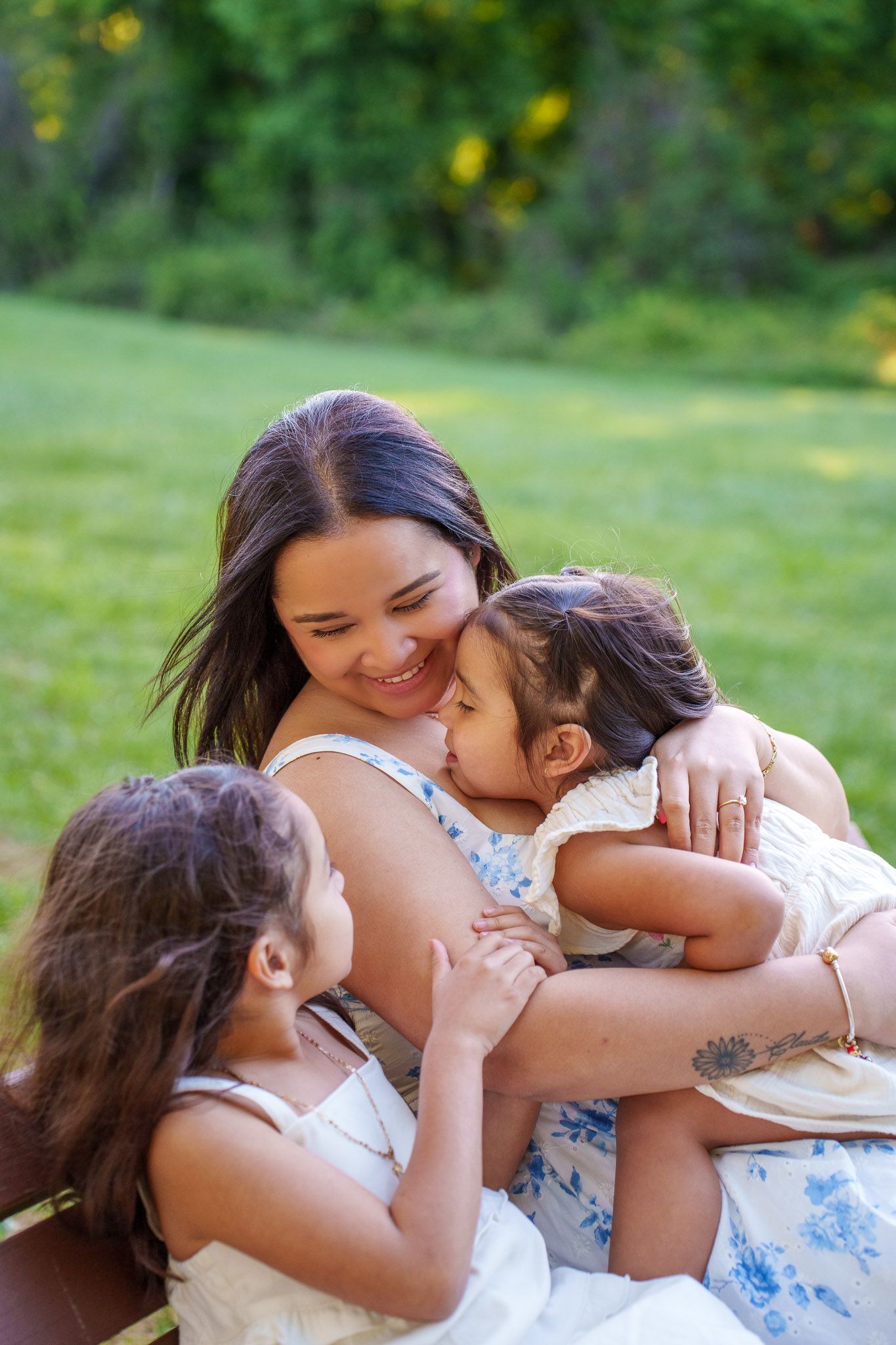 A smiling mother sits outdoors on a bench hugging her two young daughters in a lush green park.