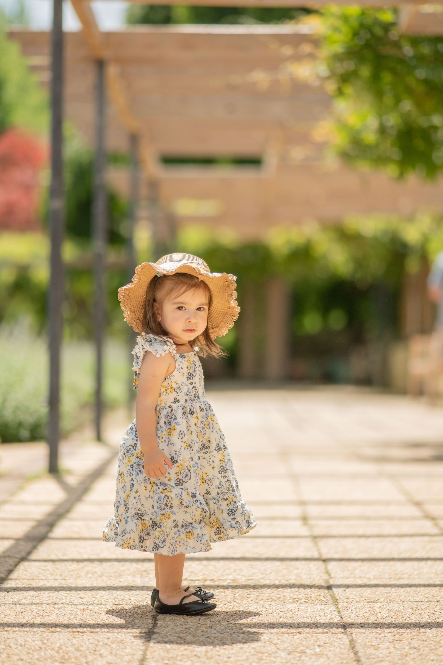 A toddler in a floral dress and straw hat plays on a stone patio in bright sunlight for portrait in Maryland