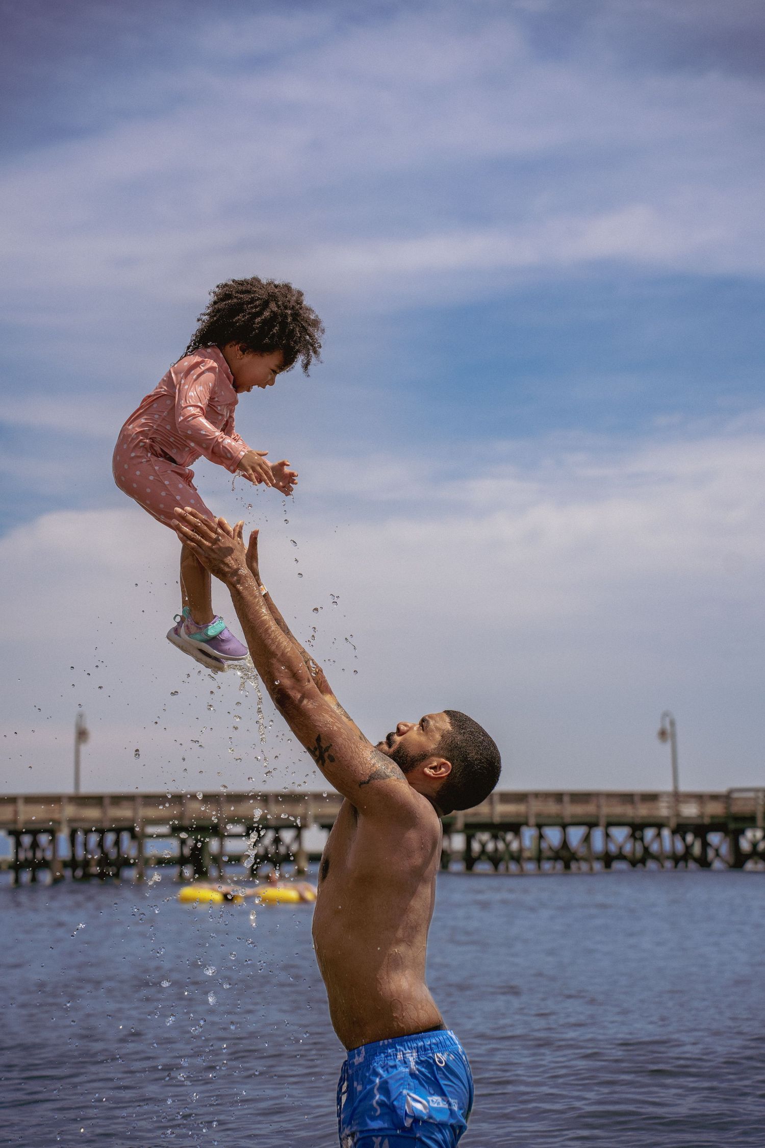 Playful moment captured of family splashing in ocean water with pier in background.