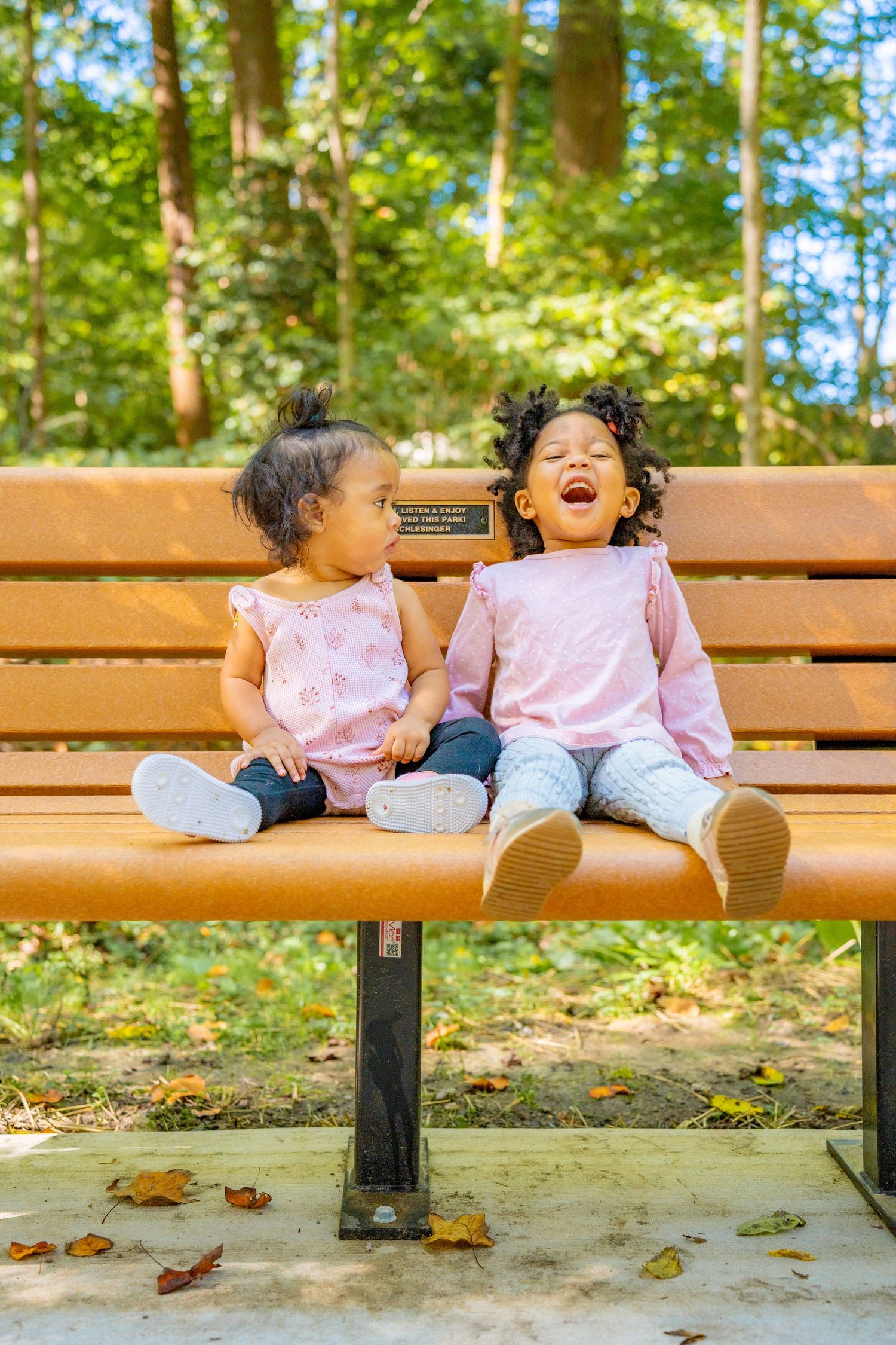 Two small children sit together on a wooden park bench surrounded by fallen autumn leaves on the ground.