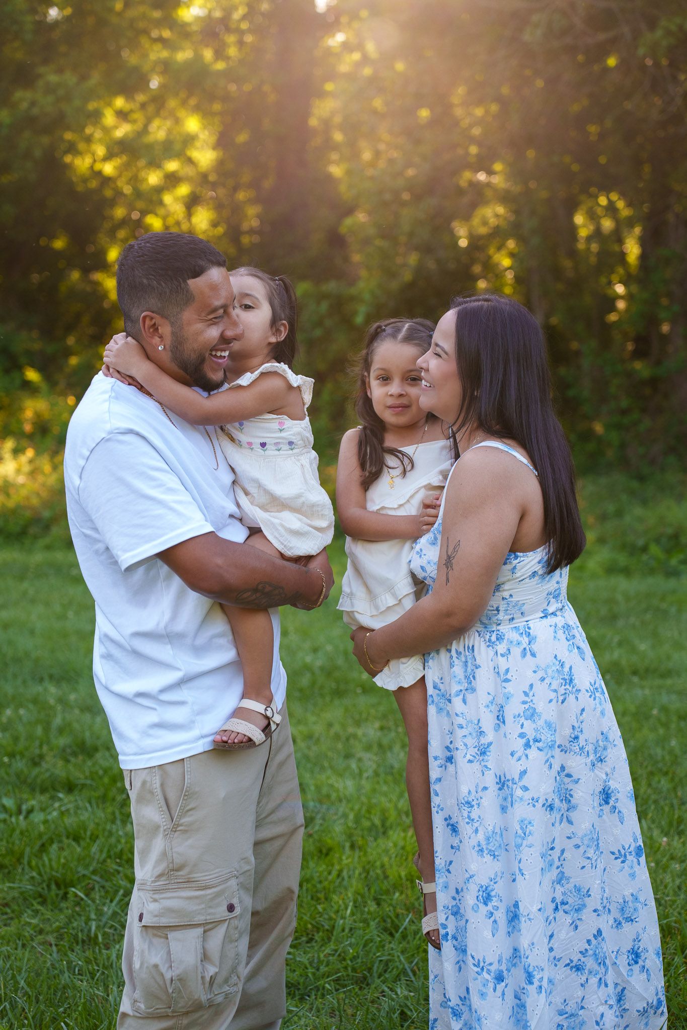 Happy family of four enjoying a golden hour outdoor photo session in a lush green park, parents holding two young daughters.