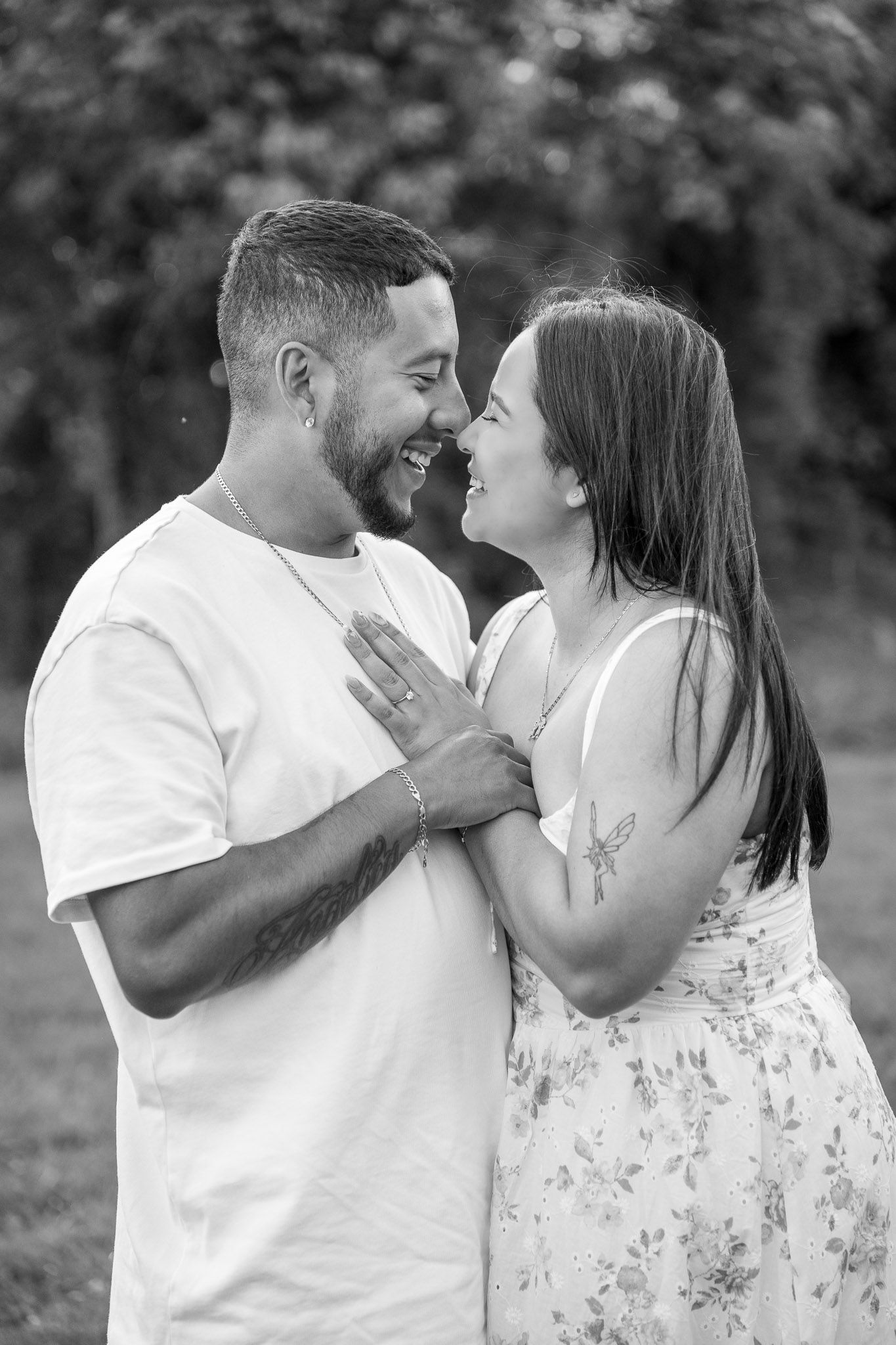 Black and white photo of a laughing couple embracing outdoors, woman placing hands on mans chest, trees in background.