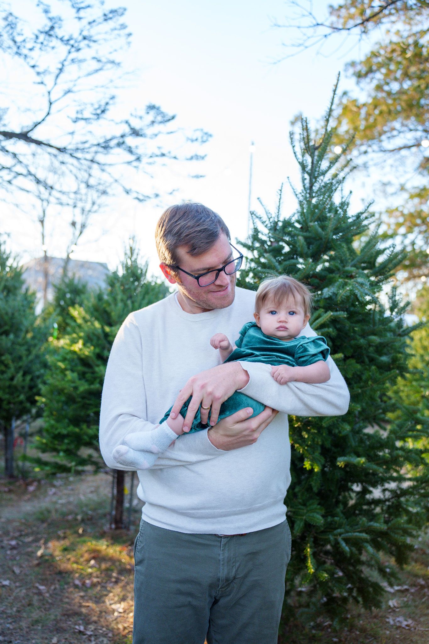 Father in white sweater holding baby in green outfit at Christmas tree farm outdoors.