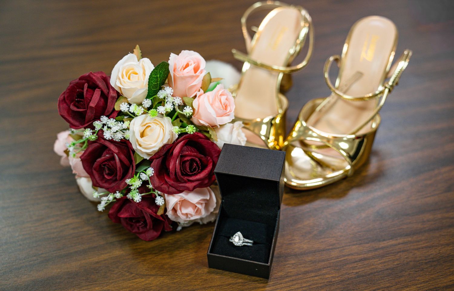 Wedding details including engagement ring in black box and pink and burgundy rose bouquet on wooden surface.