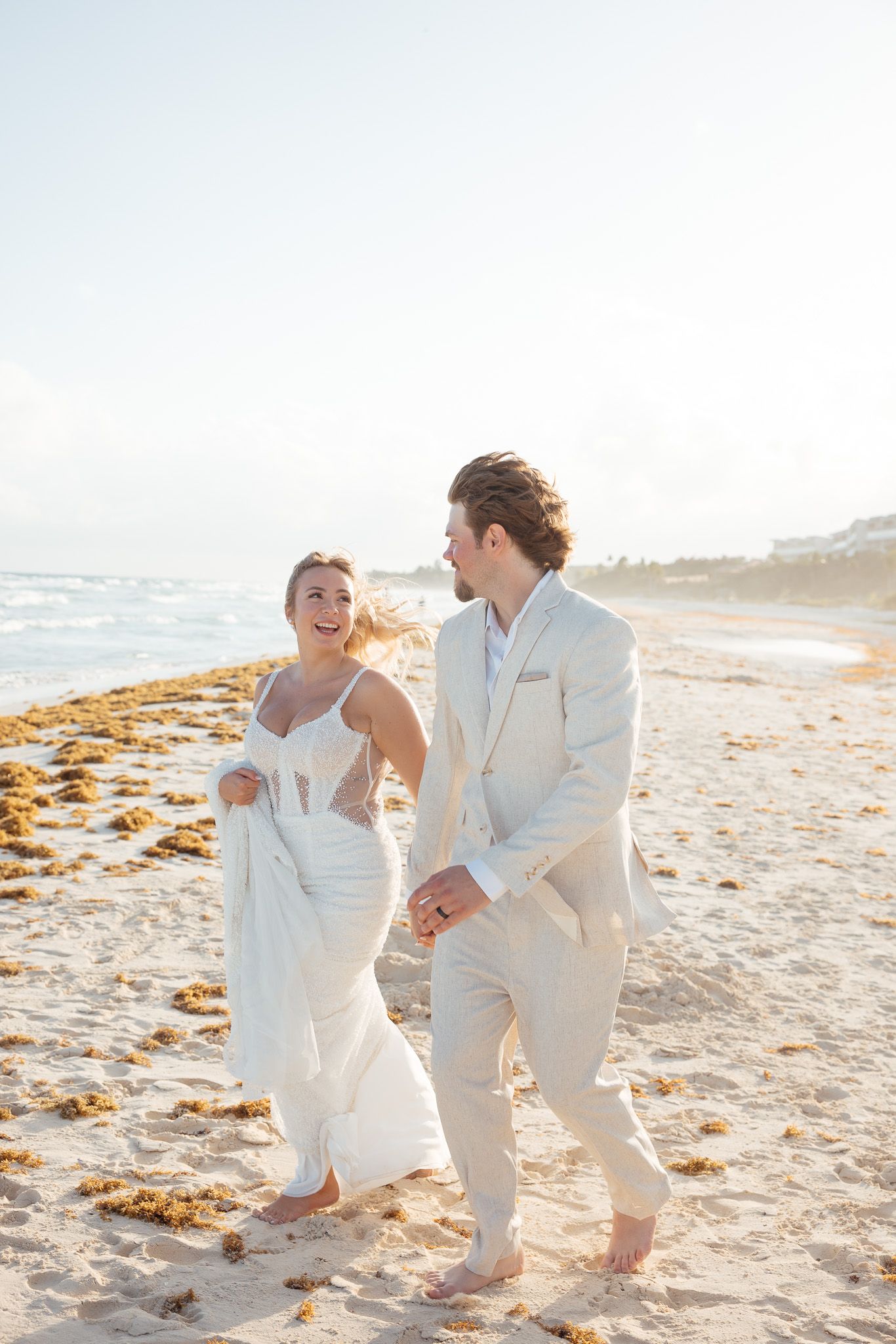 A couple in white outfits laughs and plays together on a sunny beach with seaweed washed ashore.
