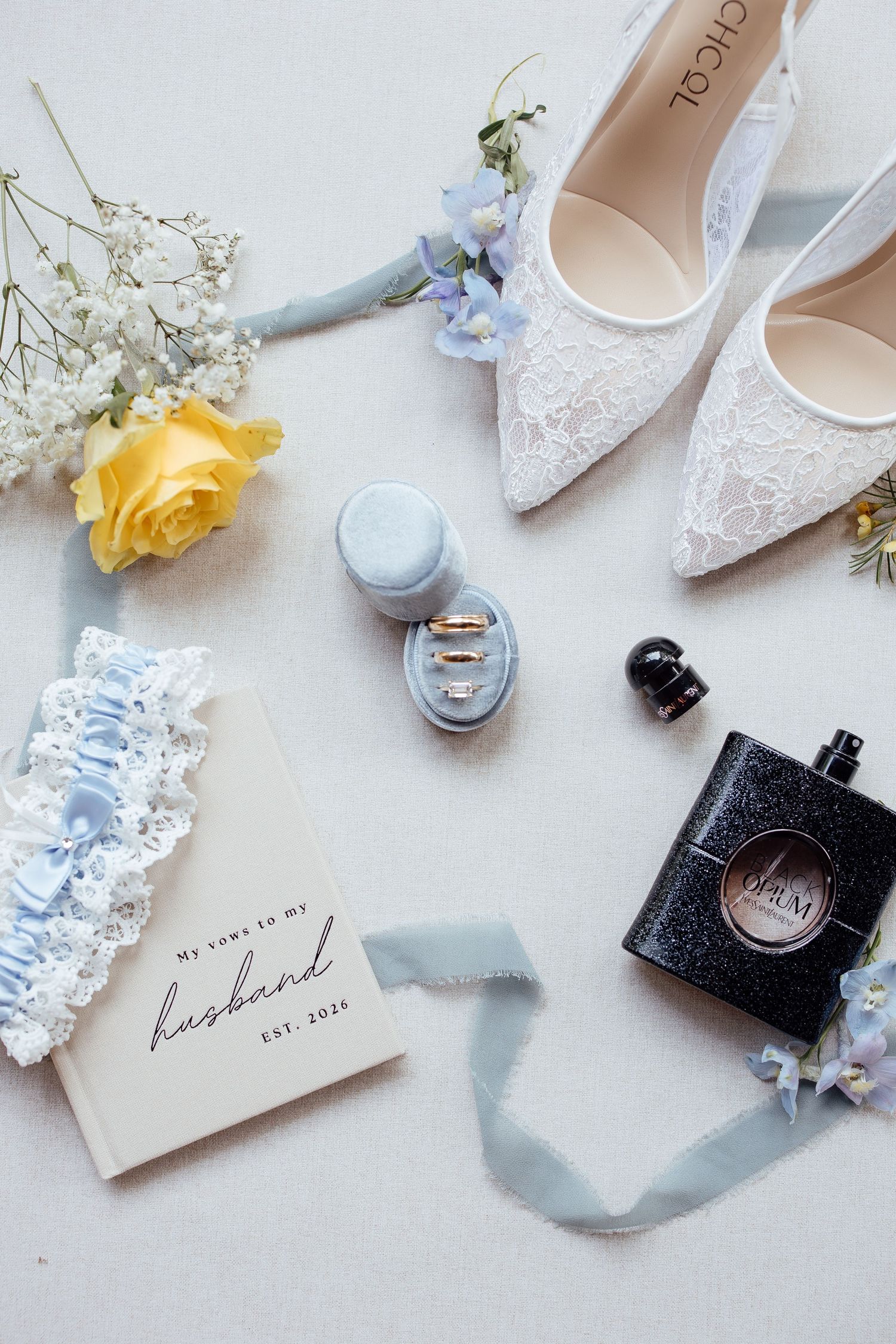 Bridal flat lay with lace heels, YSL Black Opium perfume, wedding rings, blue garter, vows book, and yellow rose.