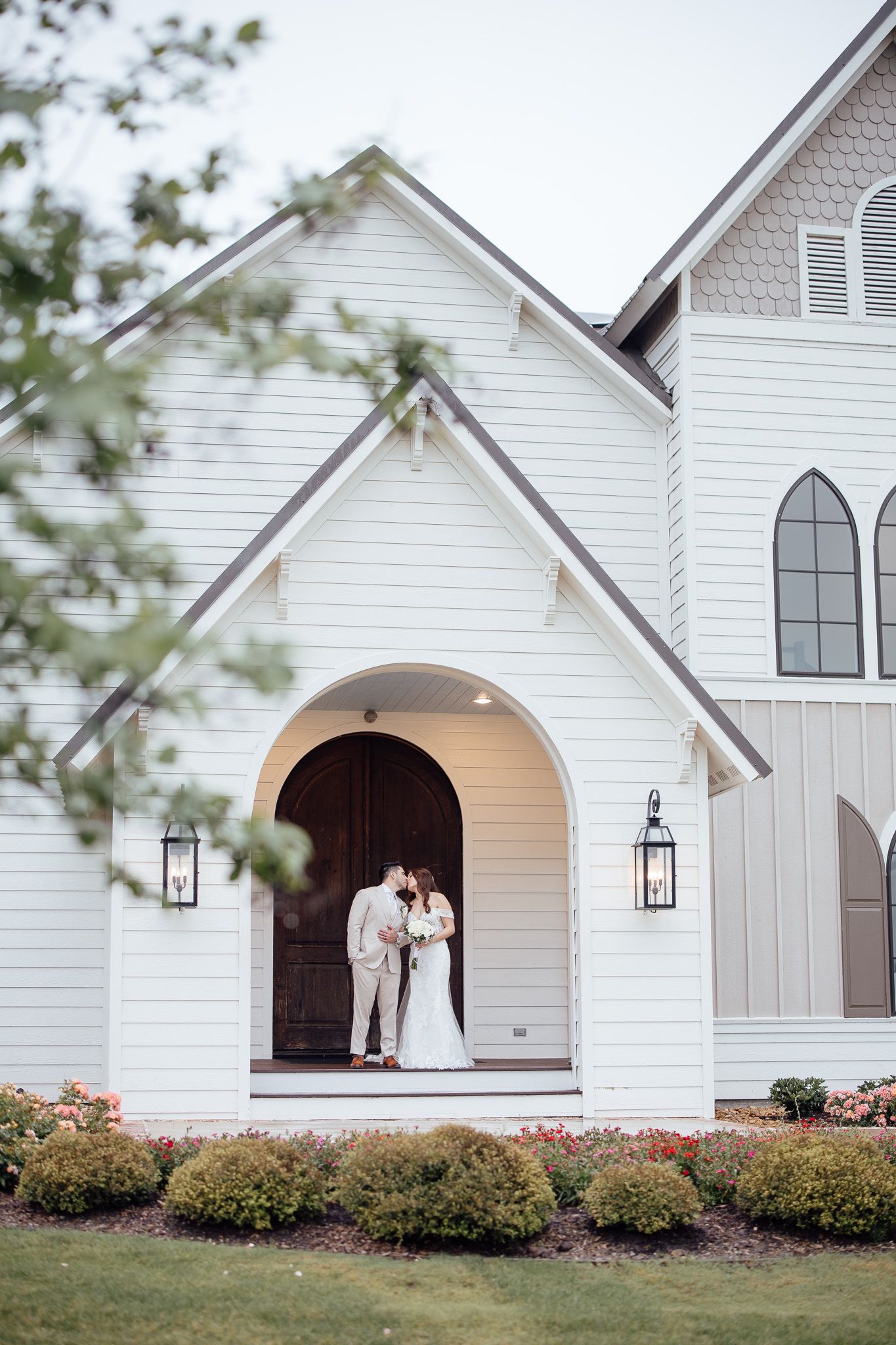 Bride and groom kissing at the entrance of a white chapel wedding venue surrounded by lush greenery.