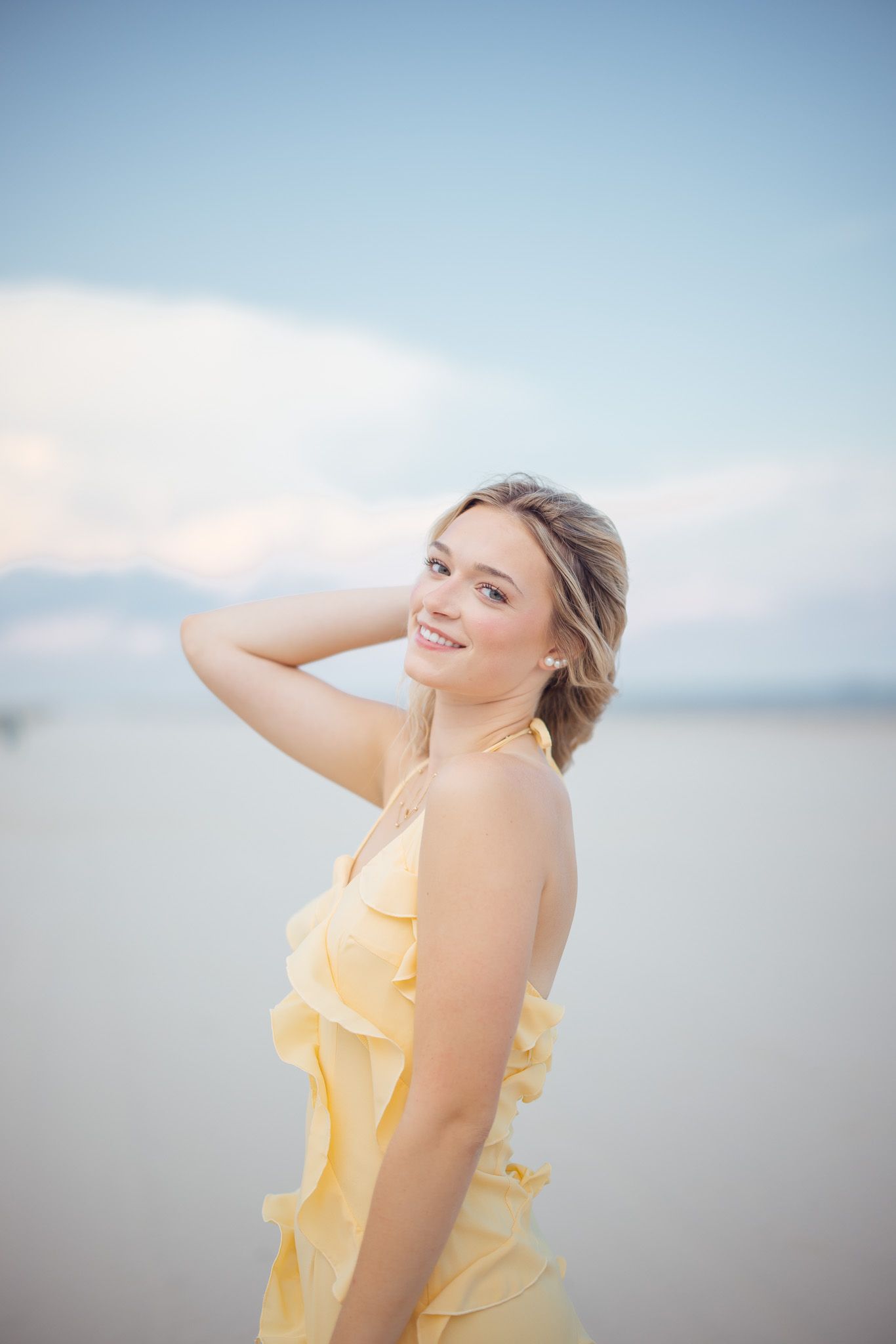 Woman in flowing yellow dress poses joyfully against soft blue sky background during outdoor photoshoot.
