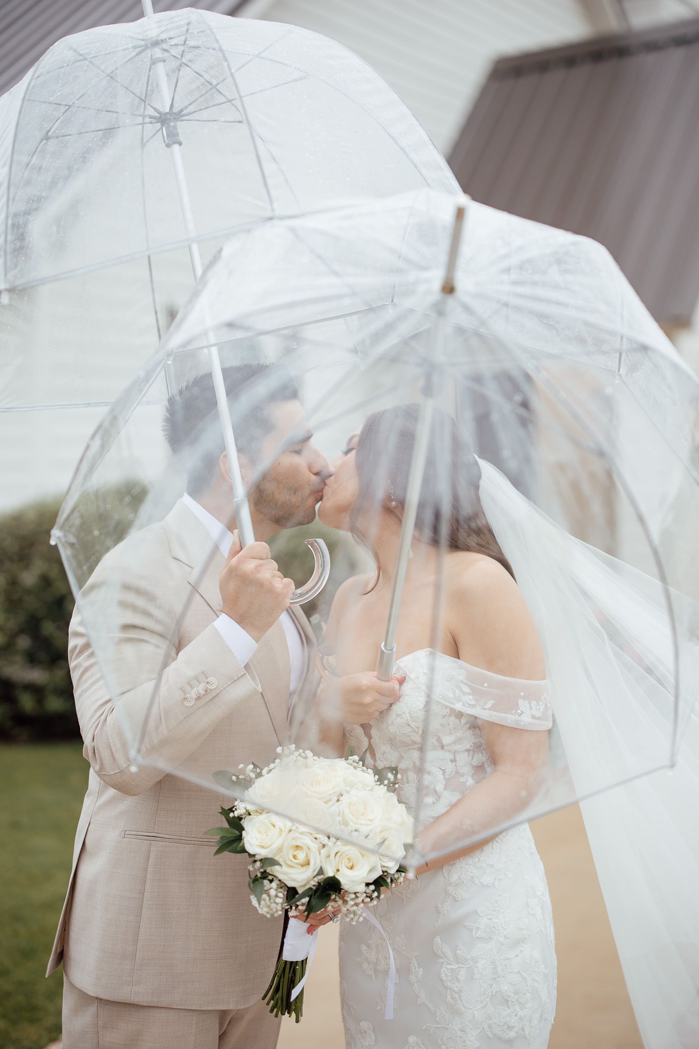 Bride and groom kissing under clear bubble umbrellas on rainy wedding day, bride holding white rose bouquet.