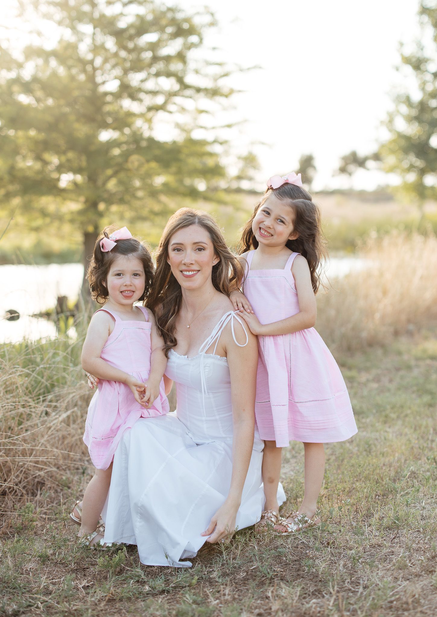 A group relaxes together on a grassy hillside wearing white and pink dresses in soft afternoon light.