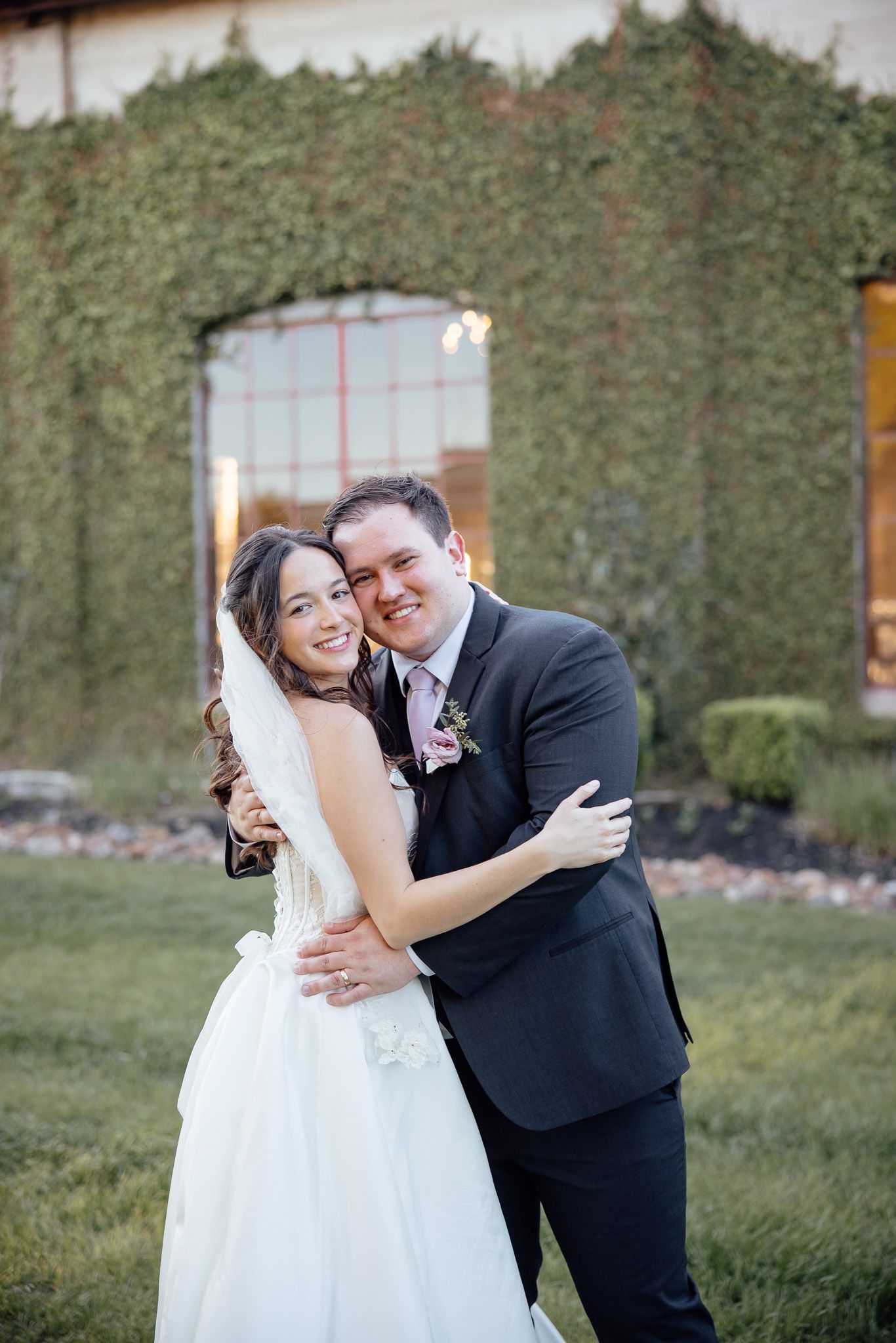 Bride and groom embracing and smiling outdoors in front of an ivy-covered building on their wedding day.