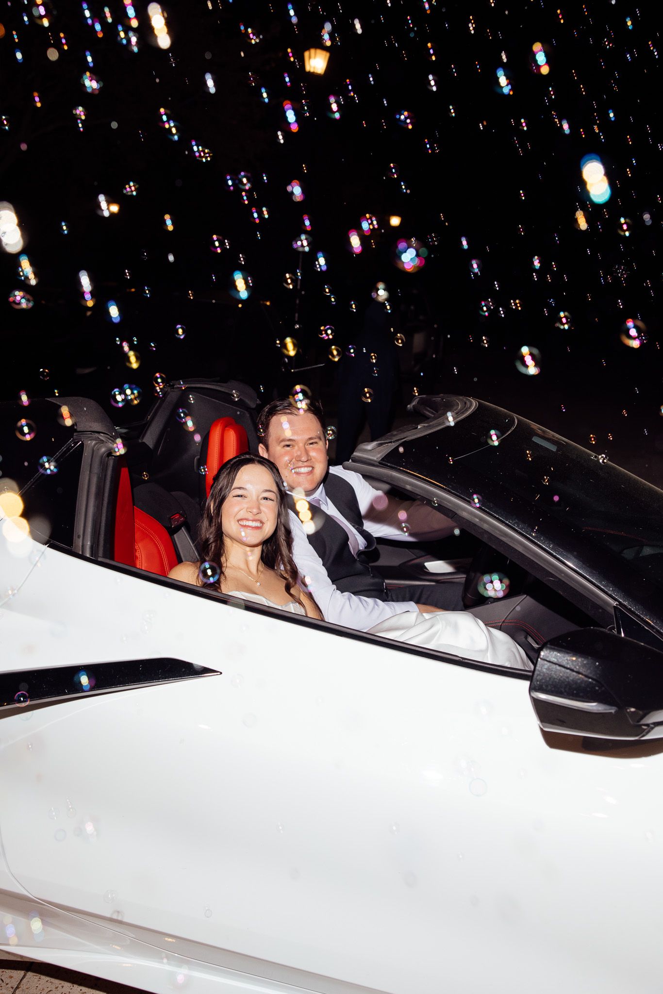 Bride and groom smiling in a white convertible car at night surrounded by colorful bokeh bubbles.