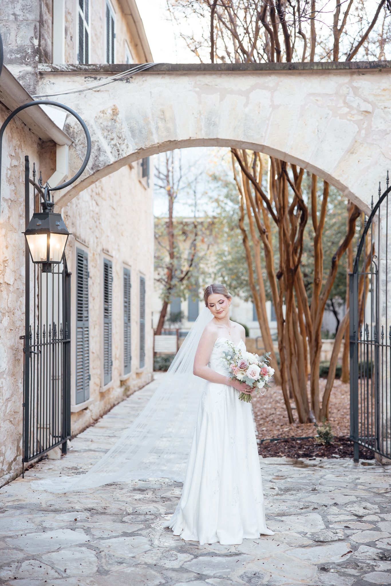 A bride in a flowing white gown and veil stands under a stone archway with crepe myrtle trees on a cobblestone path.