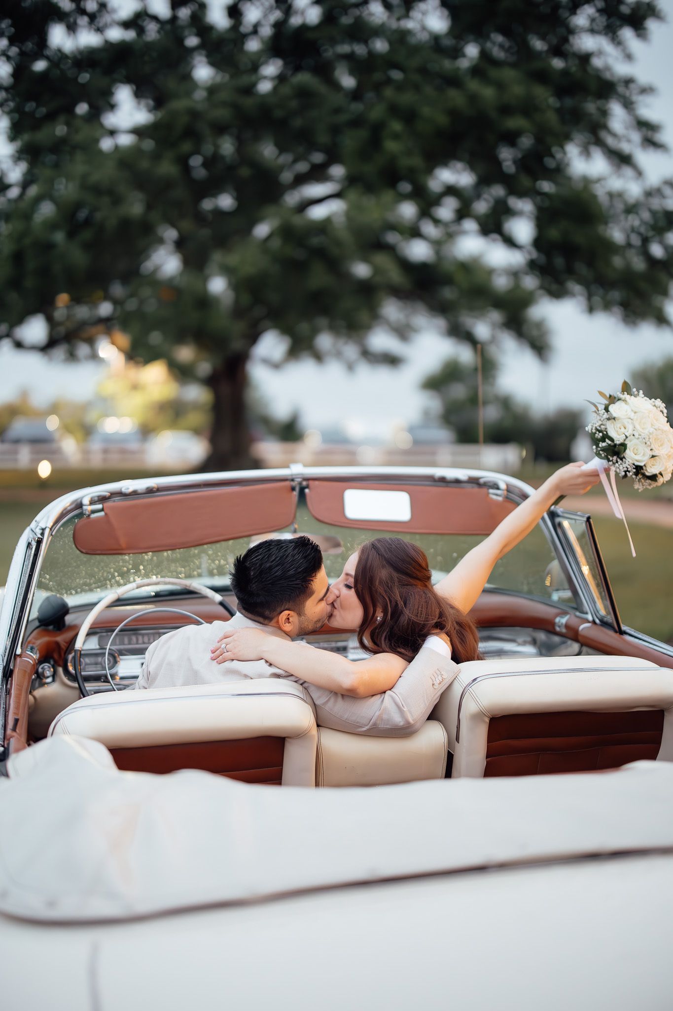 Newlywed couple kissing in a vintage white convertible car, bride holding bouquet aloft on their wedding day.