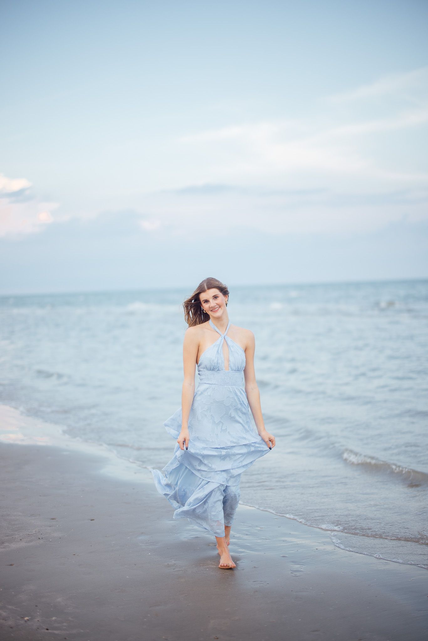 A woman in a flowing light blue dress walks barefoot along the beach at sunset with the ocean in the background.