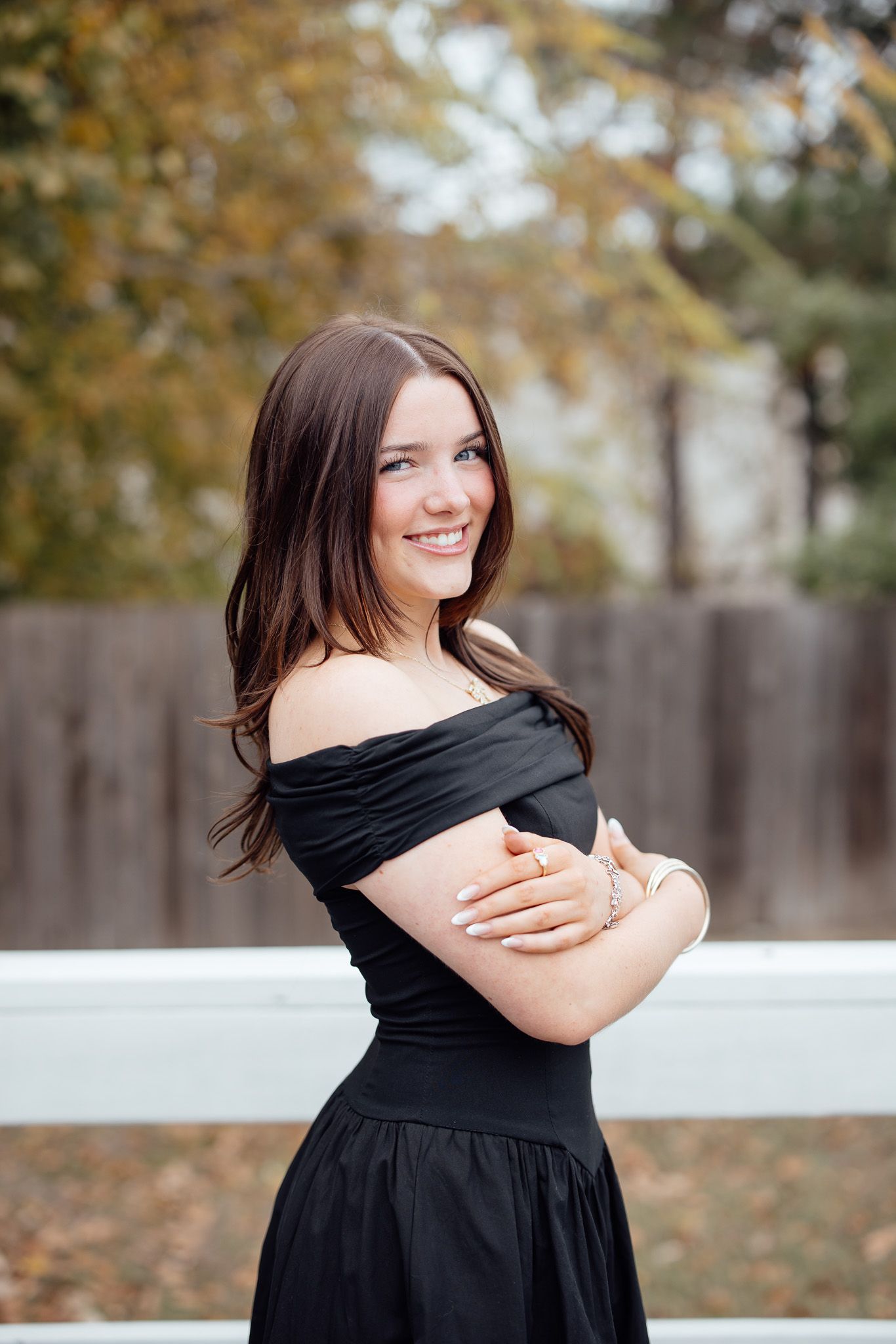 A person in a black off-shoulder dress poses outdoors against a white fence with autumn foliage in the background.