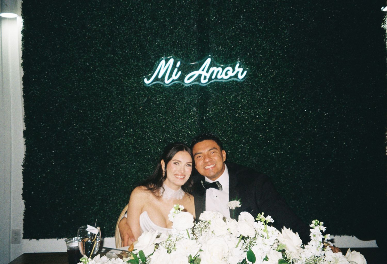Couple smiles at sweetheart table with white floral arrangements beneath a glowing Mi Amor neon sign on greenery wall.