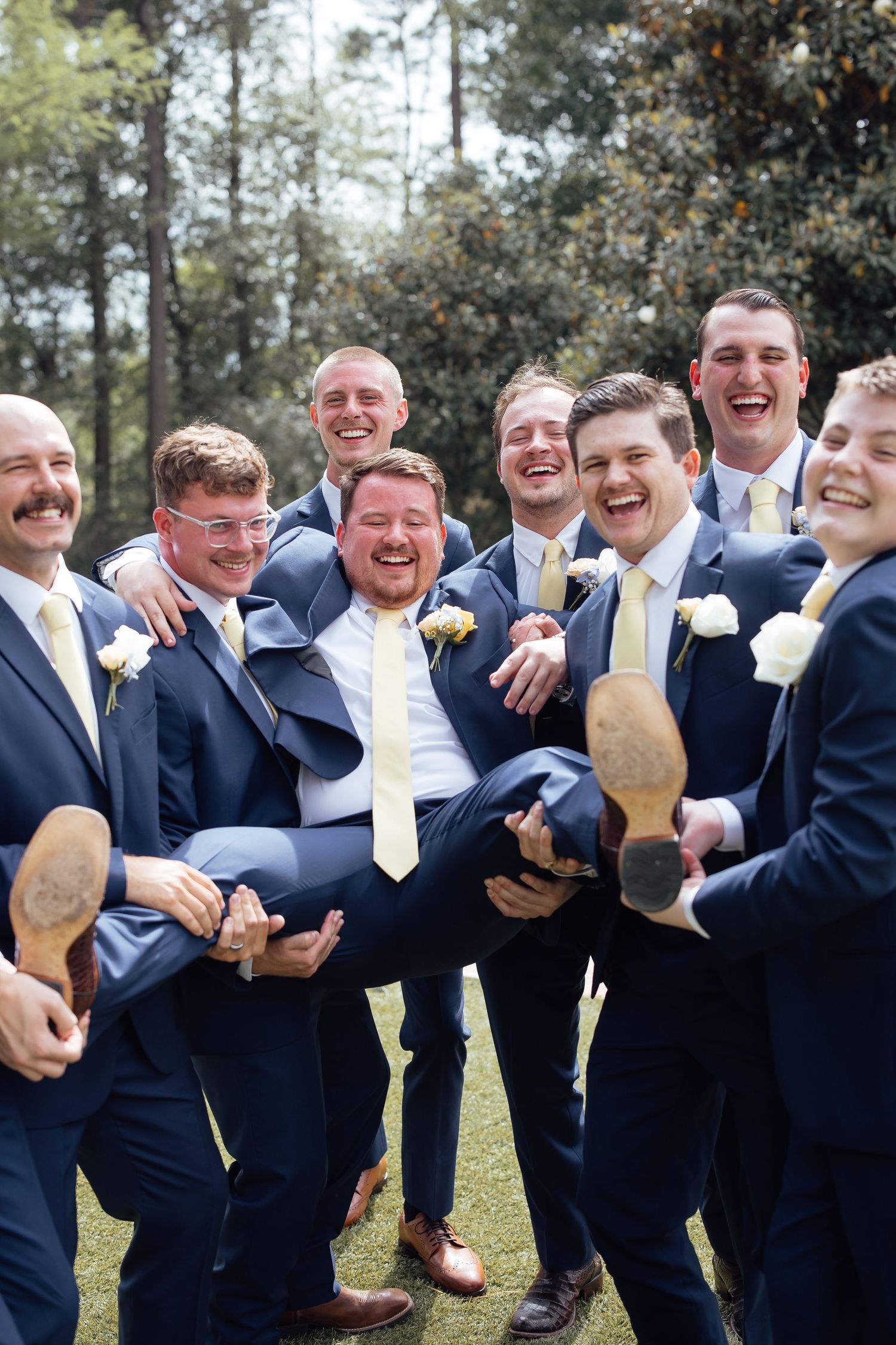 Groomsmen in navy suits and yellow ties laughing while lifting the groom during a fun wedding party photo outdoors.