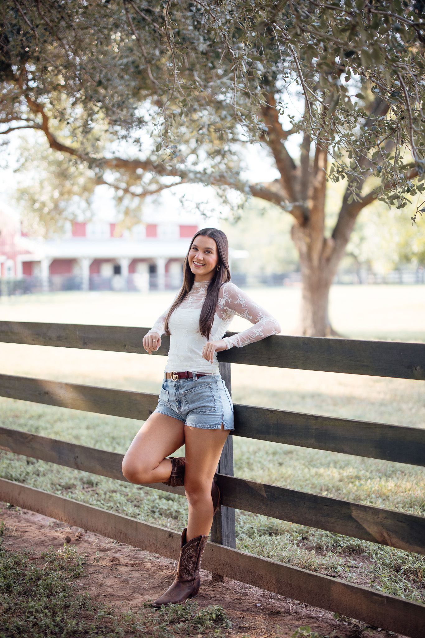 A person in denim shorts and cowboy boots poses casually on a wooden fence in a rustic outdoor setting.