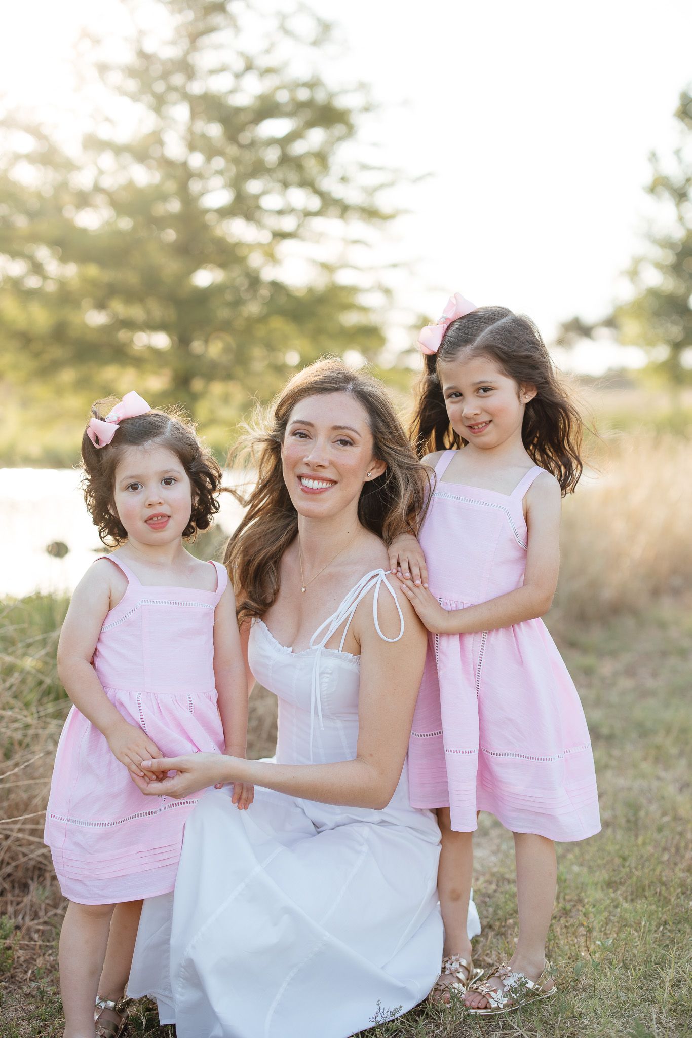 A bride in a white dress shares joyful moments with young flower girls wearing pink dresses in an outdoor field at sunset.