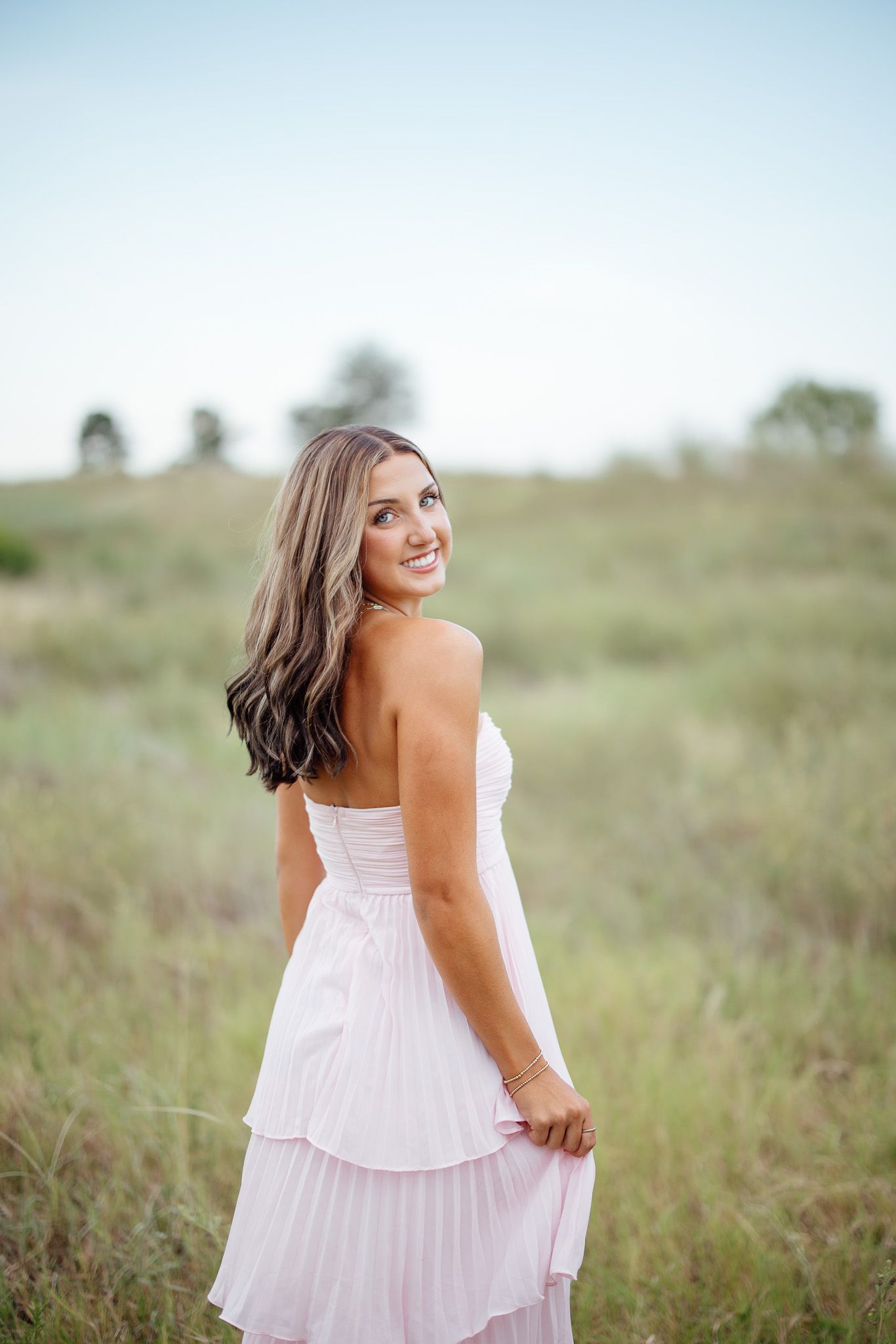 A woman in a flowing pink dress stands in a grassy field on a bright summer day.