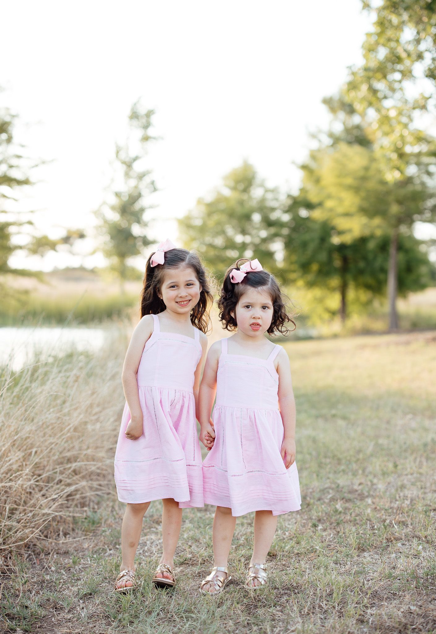 Two children wearing matching pink sleeveless dresses standing together in a grassy outdoor setting on a sunny day.