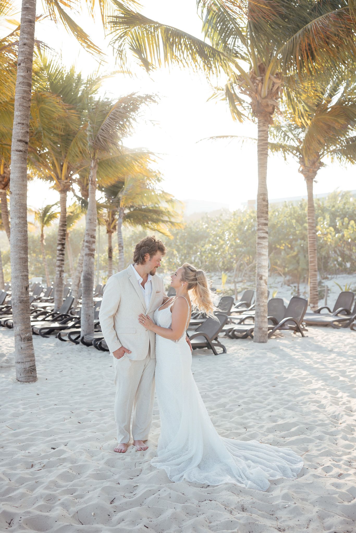 A romantic beach wedding photoshoot with palm trees and lounge chairs lining the sandy shore at sunset.