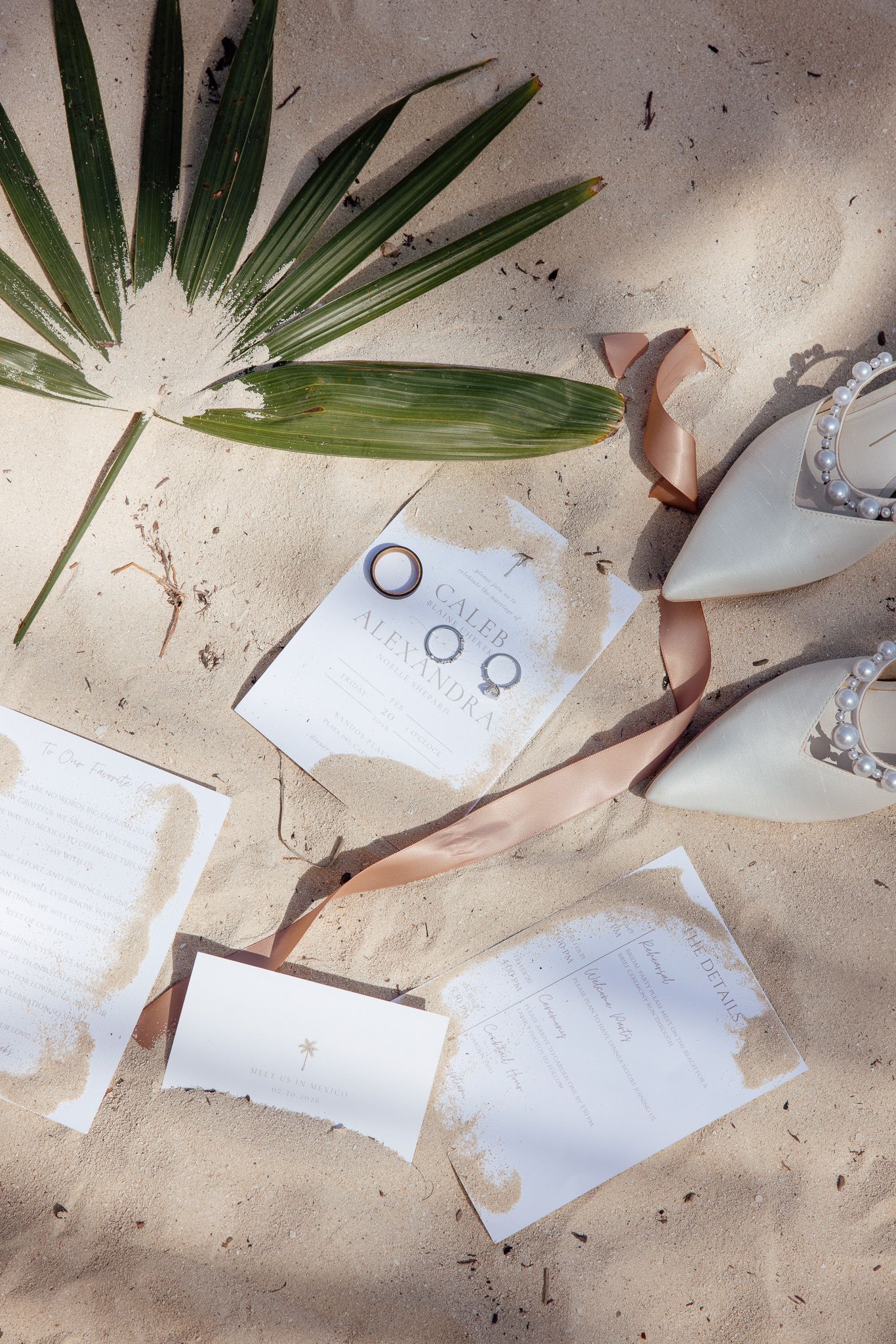 Wedding invitations with palm leaves and ribbon scattered on a beige textured surface create an elegant flat lay composition.