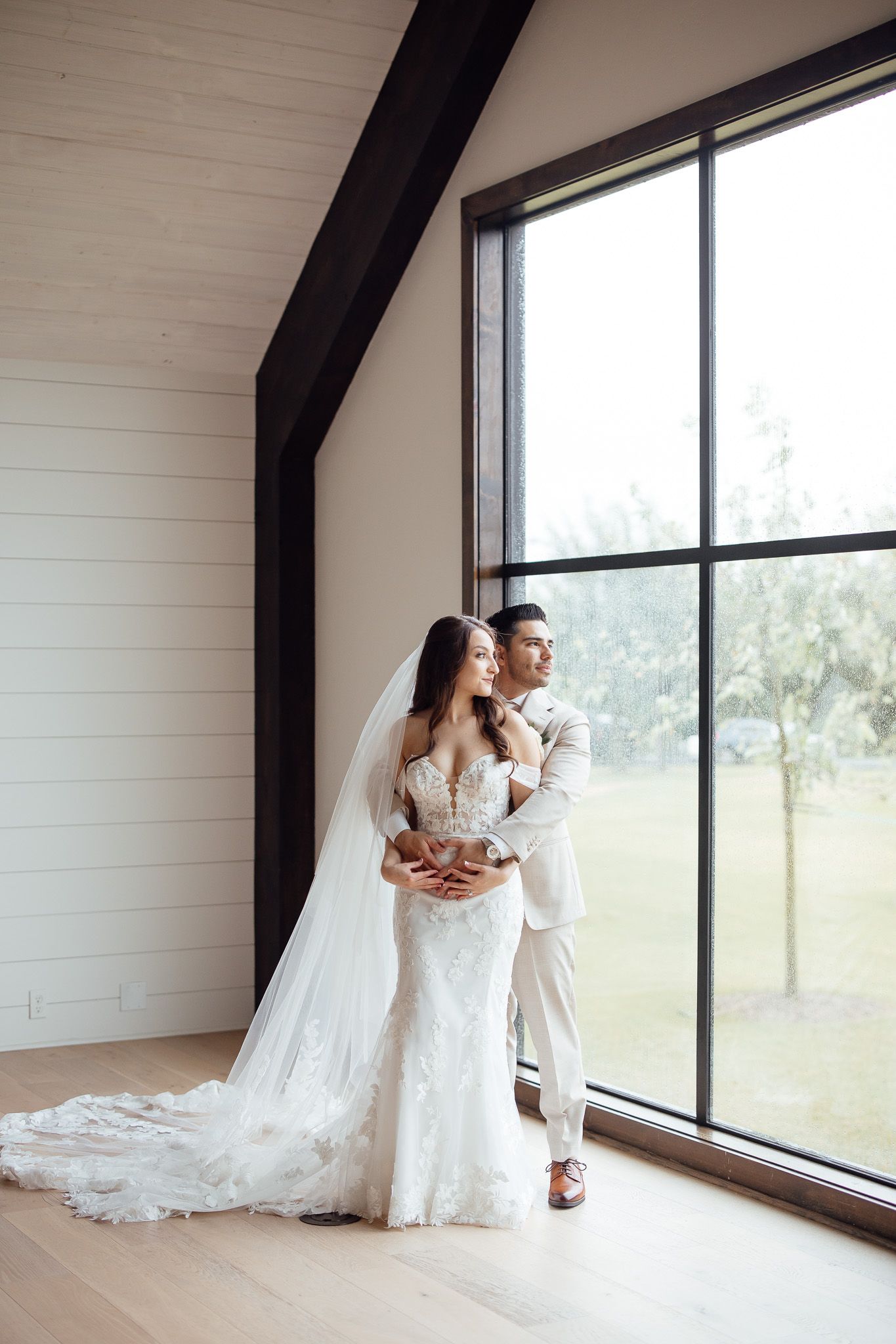 Bride and groom embrace by large window in modern barn venue, bride wearing lace mermaid gown with cathedral veil.