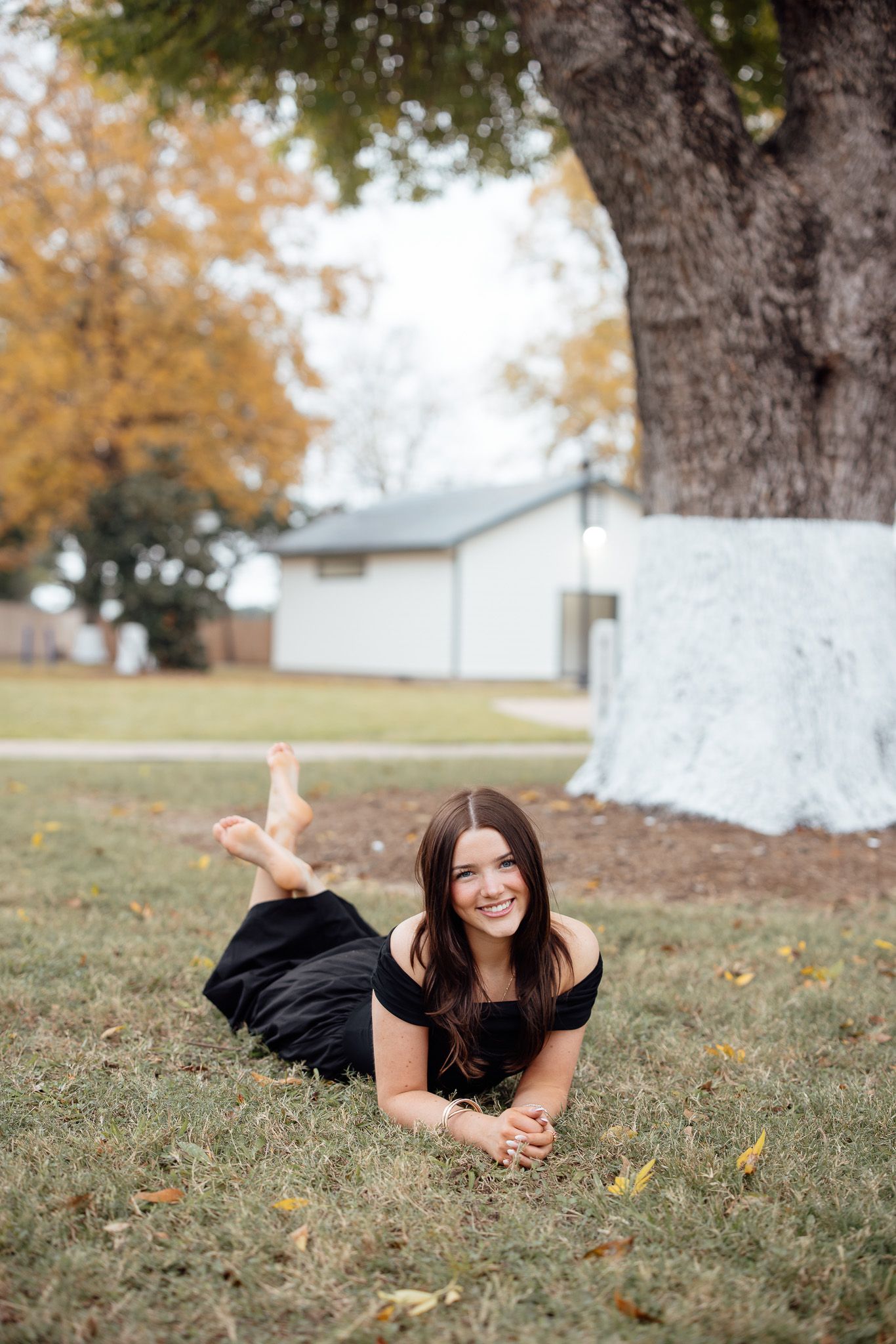 A person in a black dress lies on autumn grass beneath a large tree during an outdoor photo session.