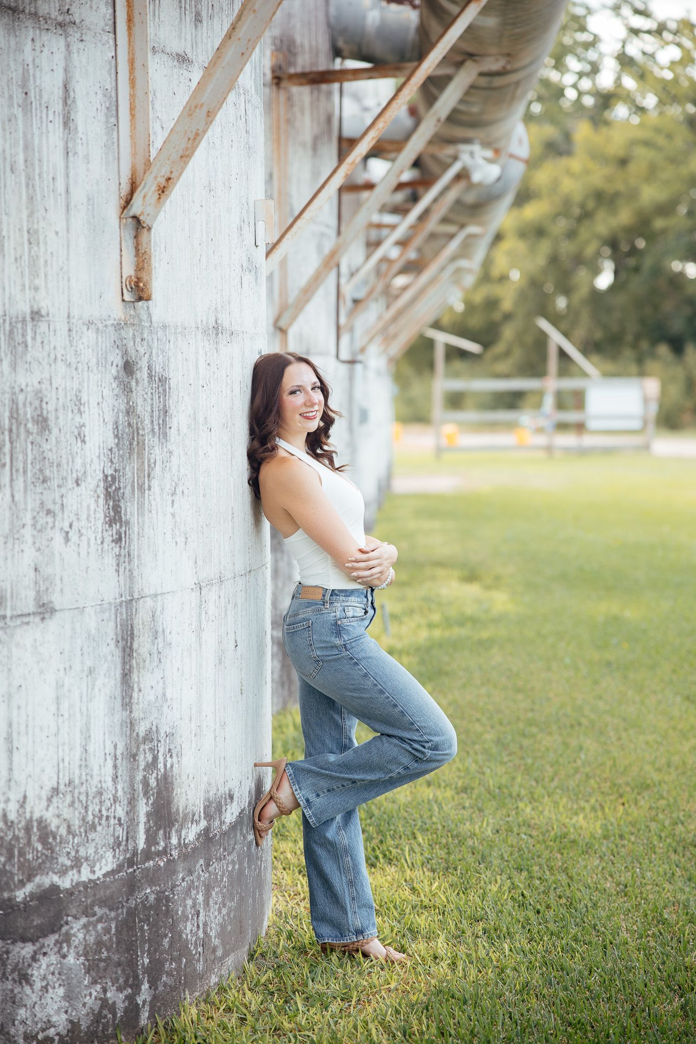 A person in overalls poses casually against a rustic white barn wall on a grassy lawn in natural lighting.