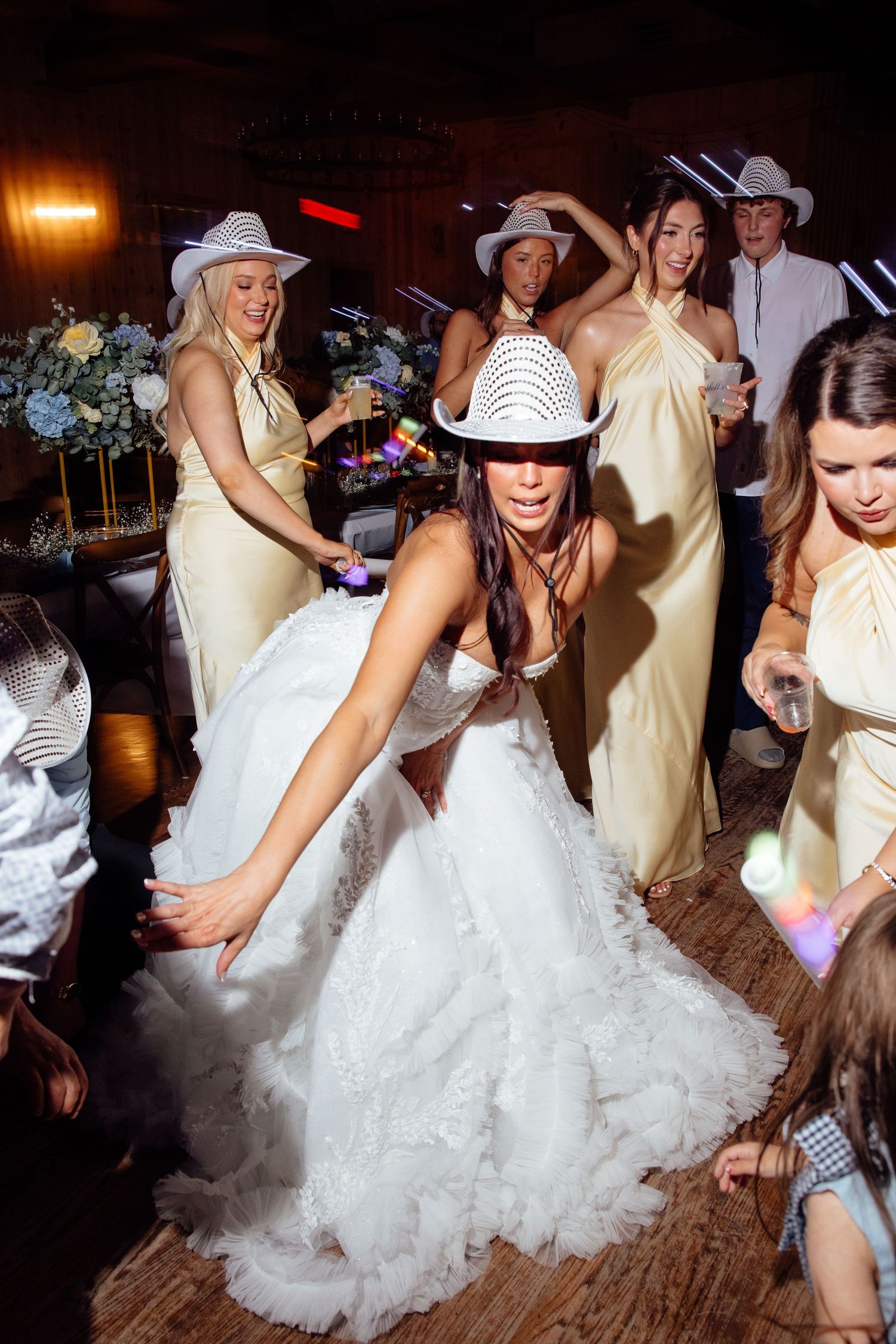 Bride in white gown and cowboy hat dancing energetically with bridesmaids in gold dresses at a lively wedding reception.