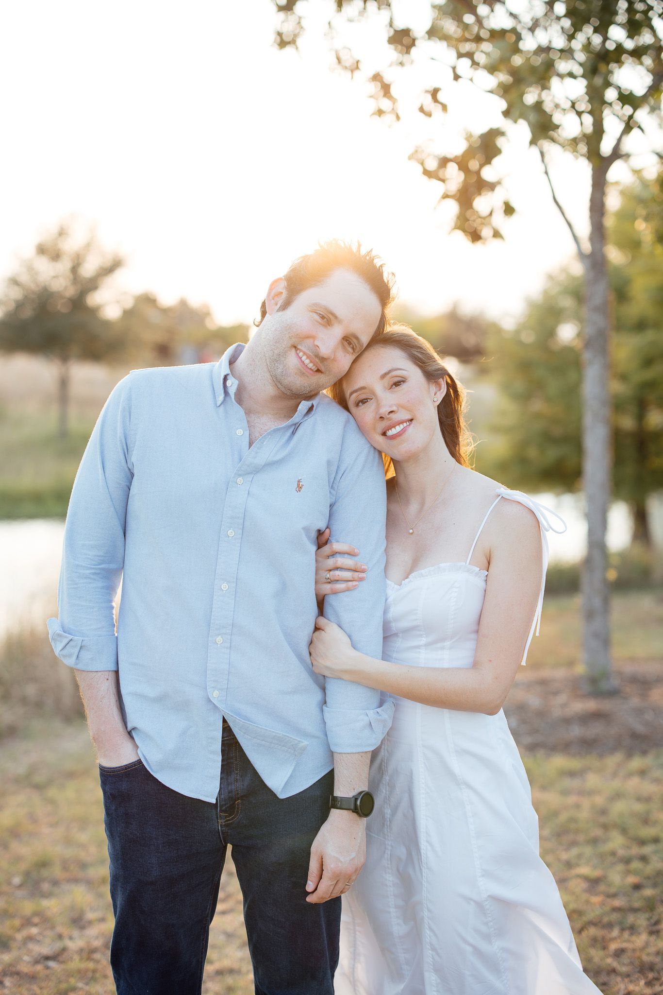 A couple in light blue shirt and white dress embrace outdoors during golden hour sunset in a park setting.