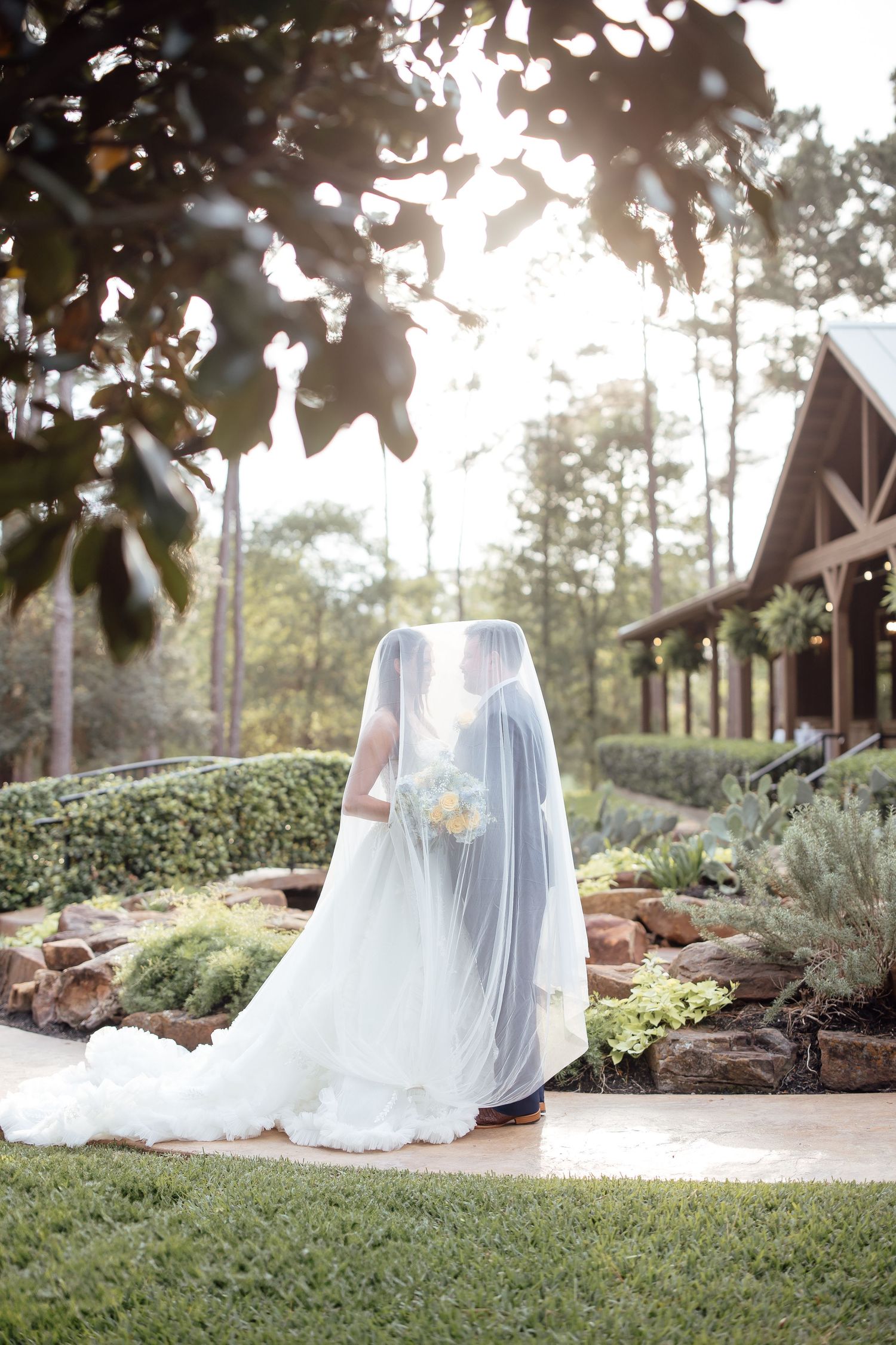Bride and groom share a kiss under a flowing veil in a lush garden setting on their wedding day.