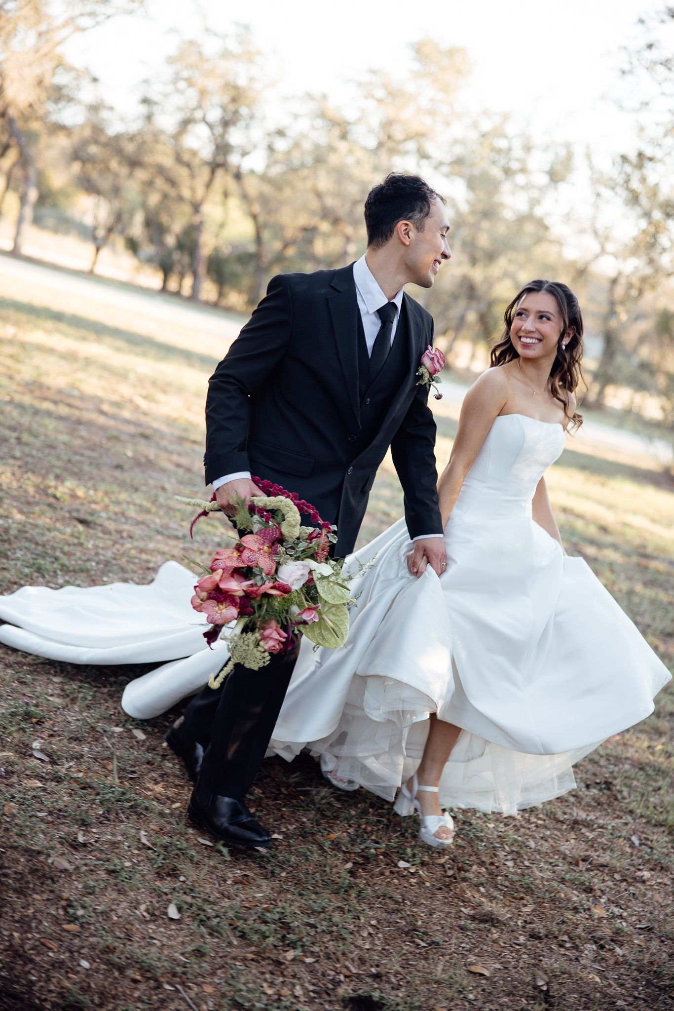 A couple in wedding attire sits playfully on autumn ground while the bride's white dress flows in the breeze.