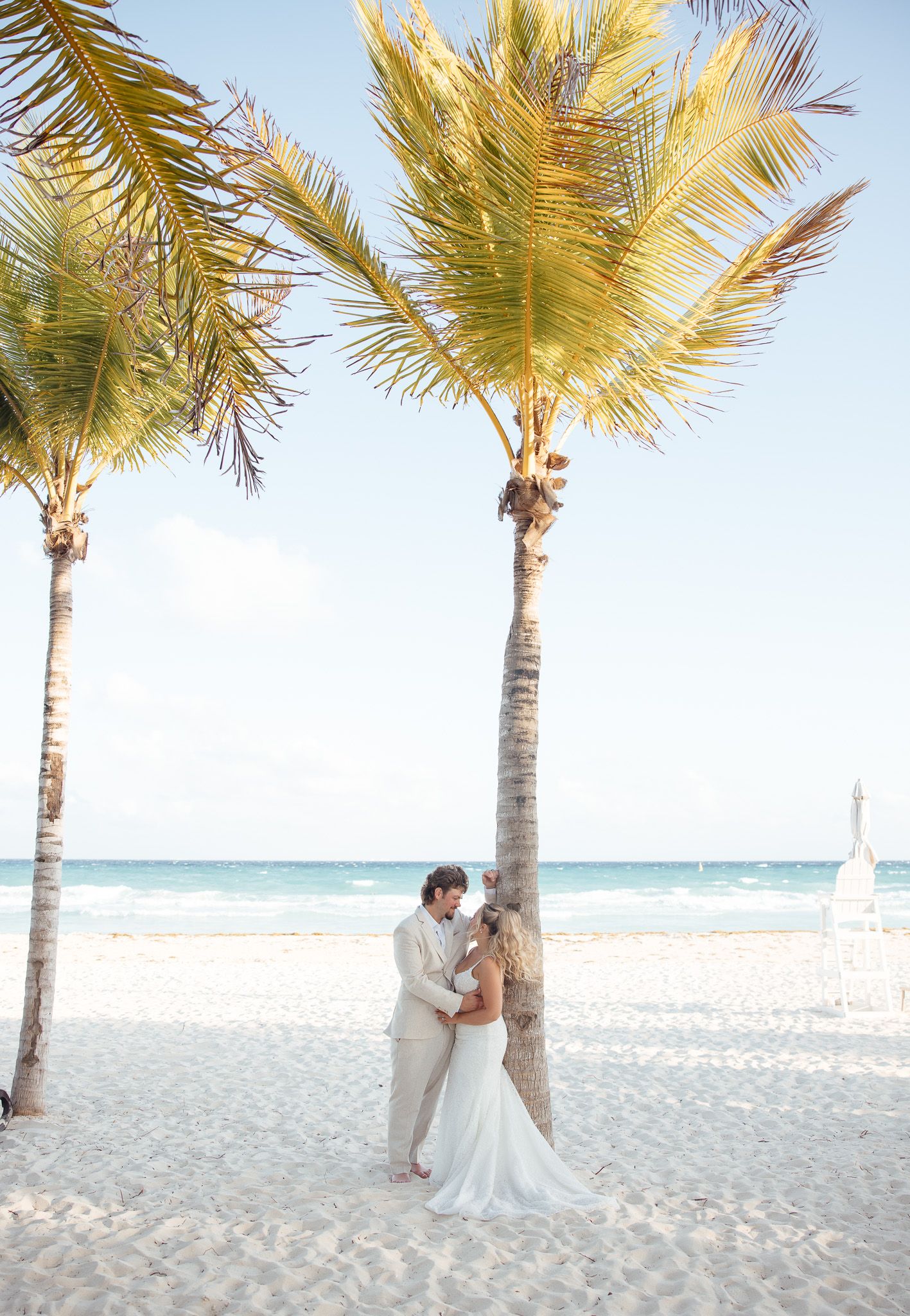Couple walking along a tropical beach with palm trees and white sand against a bright blue ocean backdrop.