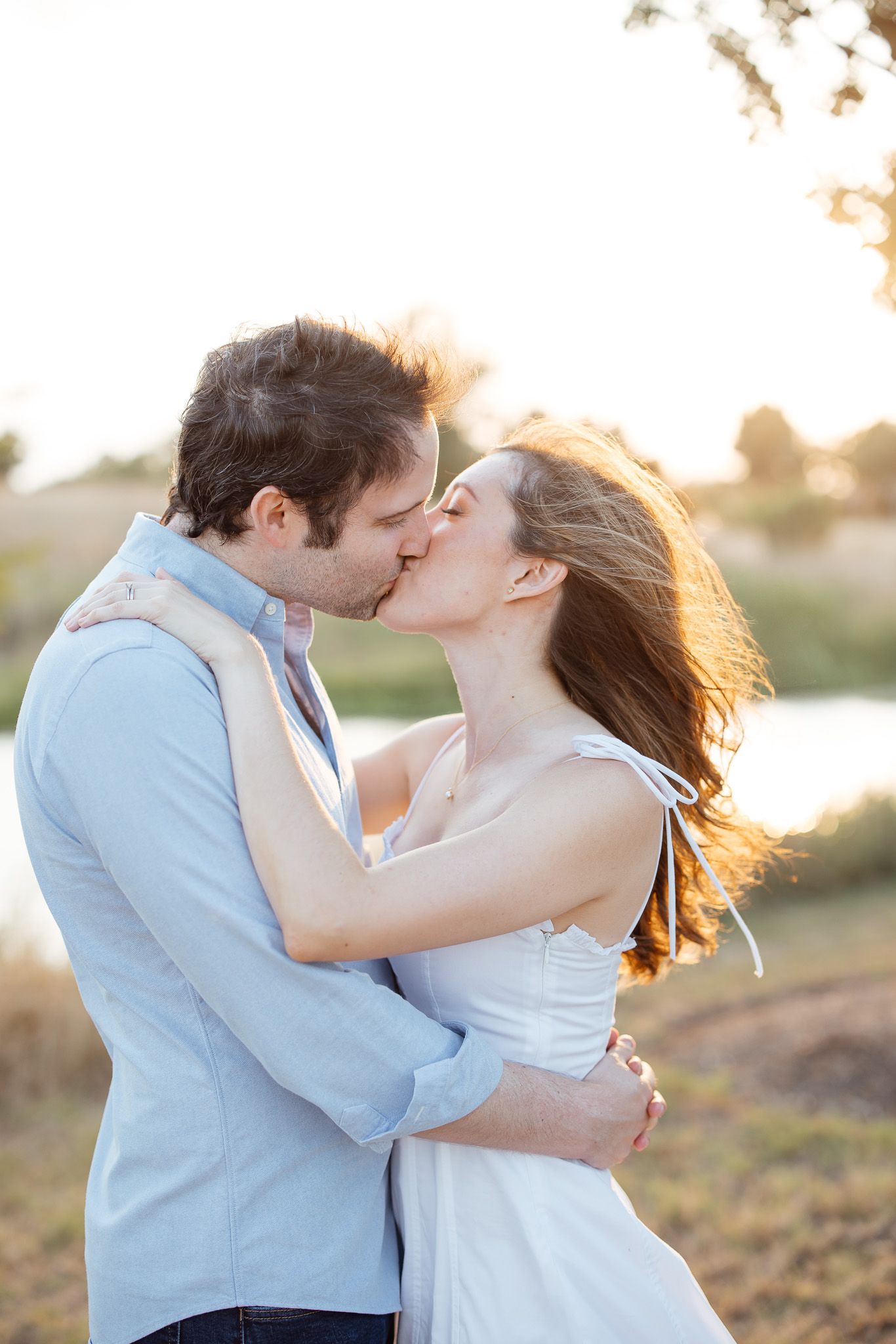 A romantic couple shares a tender kiss at sunset with golden sunlight glowing behind them by a lake.